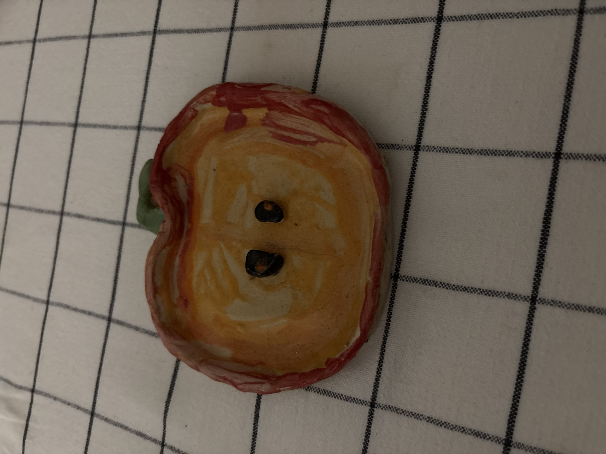 A handmade ceramic apple dish sits on a white fabric with a black grid pattern. The dish is mostly orange and red with two dark seeds in the center and a green stem. The setting appears to be indoors under artificial light. Saint Petersburg, Russia is not discernable from this image.