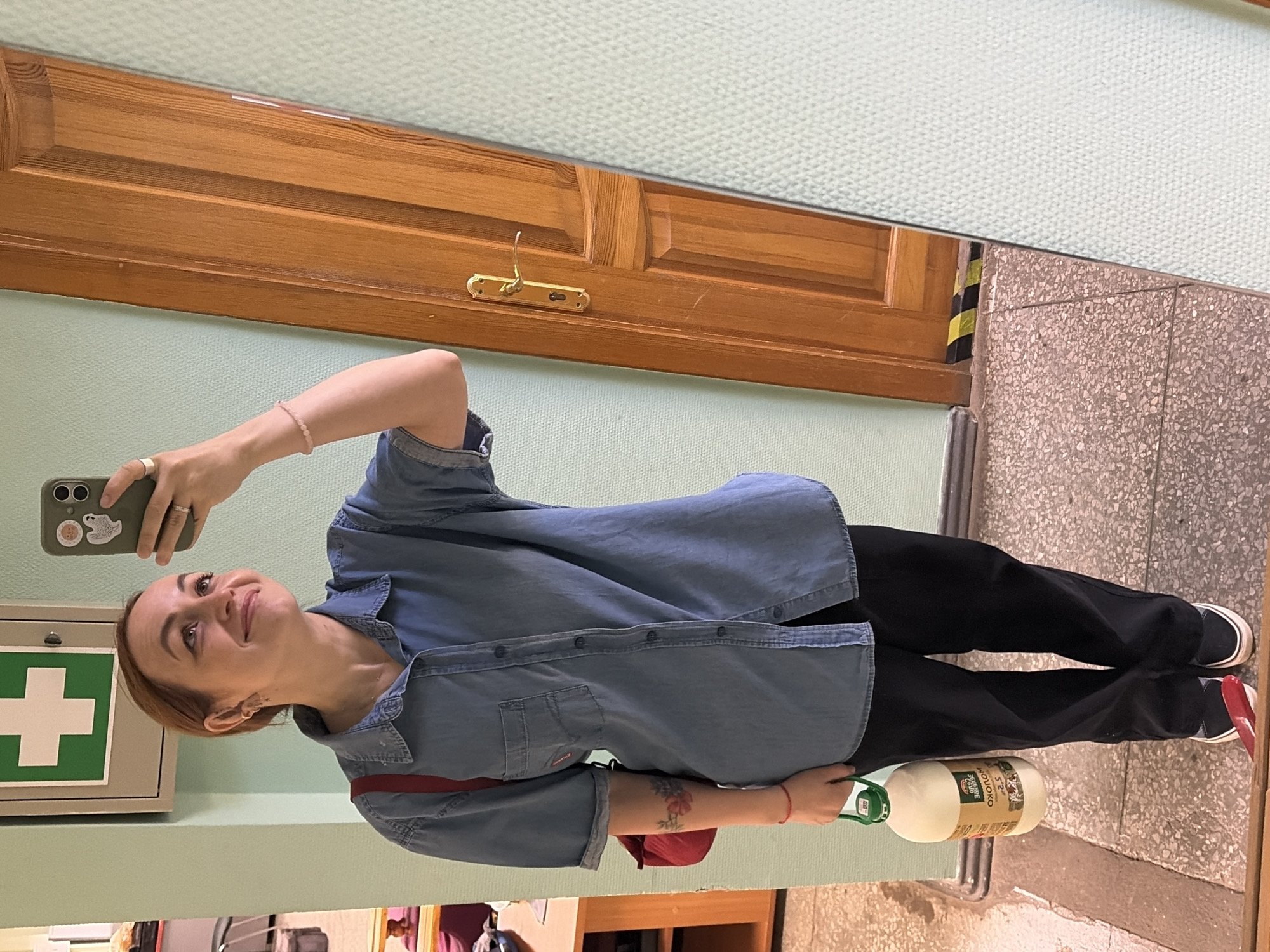A young woman with reddish-brown hair is taking a selfie in a hallway. She is wearing a denim shirt, black pants, and black sneakers with white soles. A red bag is slung over her left shoulder. In her right hand, she holds a large plastic jug of milk. A tattoo of flowers is visible on her left forearm. She is standing in front of a pale green wall and a wooden door. To the left of the door, a first aid kit is mounted on the wall. The floor is a speckled gray and pink tile. The scene appears to be inside a building in the city of Chaikovsky, Russia.