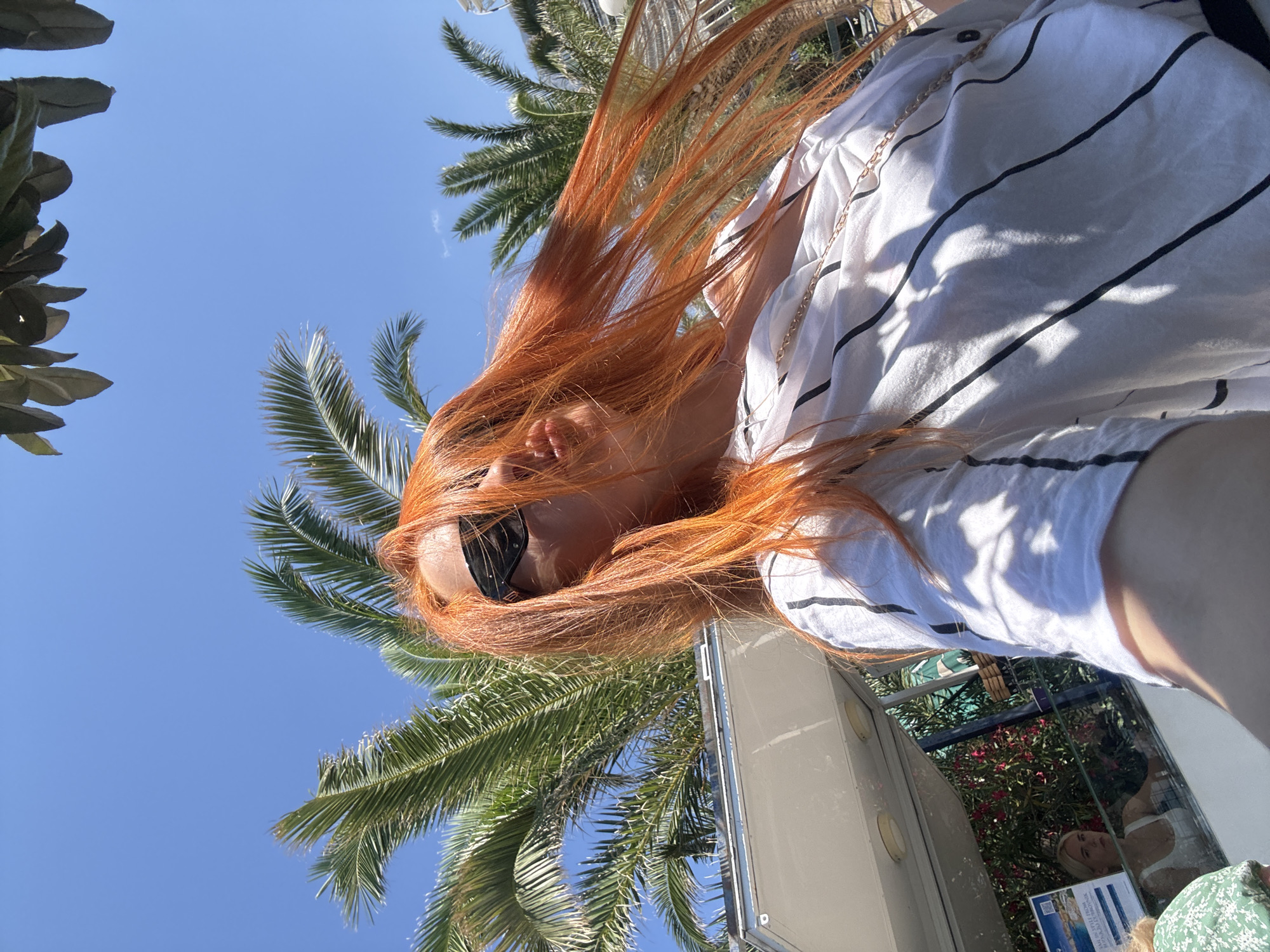 The image shows a close-up, low-angle shot of a person with vibrant red hair blowing in the wind, set against a bright blue sky and palm trees. The person is wearing sunglasses and a white shirt with black stripes, adorned with a gold chain. Sunlight casts shadows on the shirt. In the background, a modern structure, possibly a building or a boat, is visible. A reflection in a glass surface shows a woman with blonde hair, a white top, and a colorful patterned garment. The scene evokes a feeling of a sunny day, possibly in a tropical or Mediterranean location. The location context of Budva, Montenegro, suggests a coastal setting. No specific time of day is indicated other than it being sunny, and no distinct emotions are conveyed. There is some visible text on a sign in the reflection, but it is not legible.