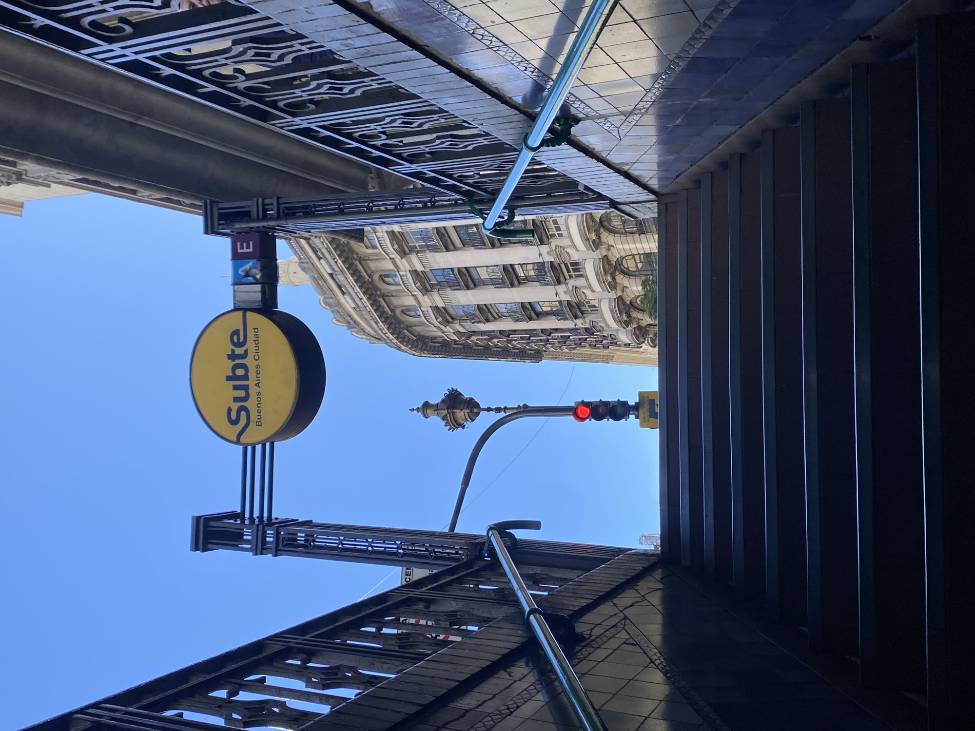 The image depicts a street view in Buenos Aires, Argentina, looking upwards from the entrance of a Subte (subway) station. The yellow Subte sign is visible, identifying the location as a subway stop. Above the subway entrance, the facade of a building with multiple windows and ornate details is visible. A streetlight with a red traffic light is positioned beyond the subway entrance. The foreground is framed by the architectural details of the subway entrance itself, including the tiling and structural supports. The sky is clear and blue.