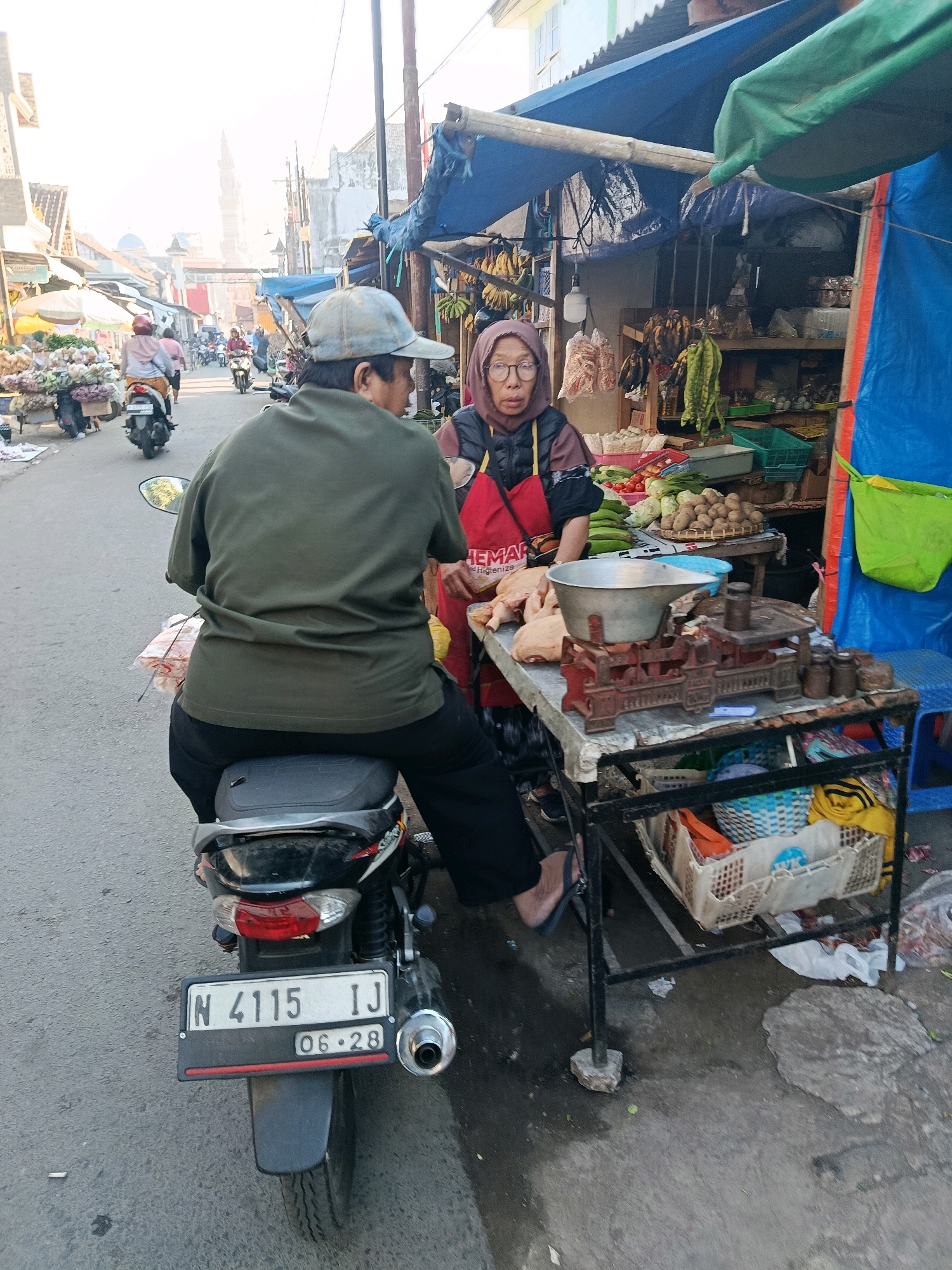 A medium shot captures a street market scene in Gondanglegi, Indonesia, focused on two individuals. In the foreground, a person wearing a green shirt and a grey baseball cap is seated on a motorcycle, facing away from the camera. To their right, a woman wearing a hijab and glasses, with a red apron over a dark jacket, stands behind a stall. The stall displays raw chicken, a vintage weighing scale with weights, and various vegetables like potatoes, tomatoes, and green beans. The background shows a bustling street with other market stalls, motorcycles, and pedestrians, suggesting a vibrant local economy. The overall lighting indicates it is daytime, likely early morning or late afternoon due to the soft, warm sunlight. The ground is paved, and there are signs of weathering and wear on the structures and pavement, typical of an outdoor market.