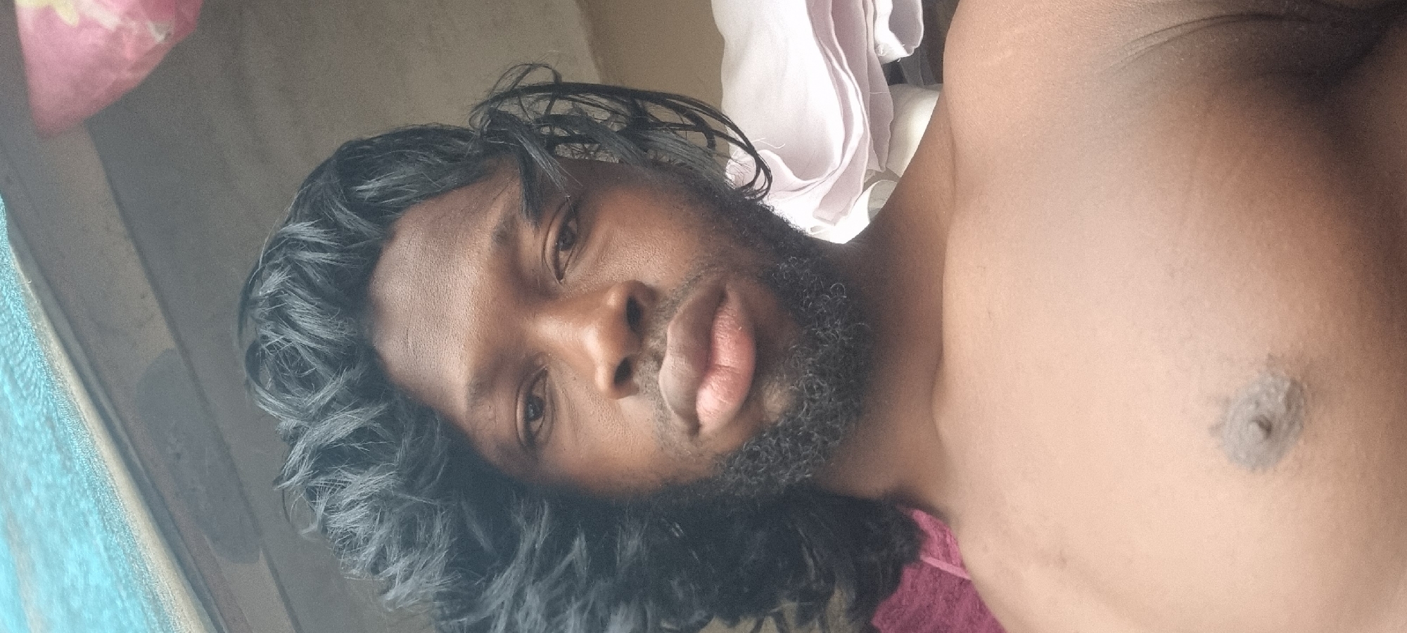 A close-up, portrait-oriented photograph shows a dark-skinned man lying down. He has black curly hair, a beard, and is shirtless, with his chest visible in the frame. The background includes a partially visible window and a bed with pink and white bedding. The location of the photograph is unknown and there is no evidence suggesting it was taken in Ikeja, Nigeria.