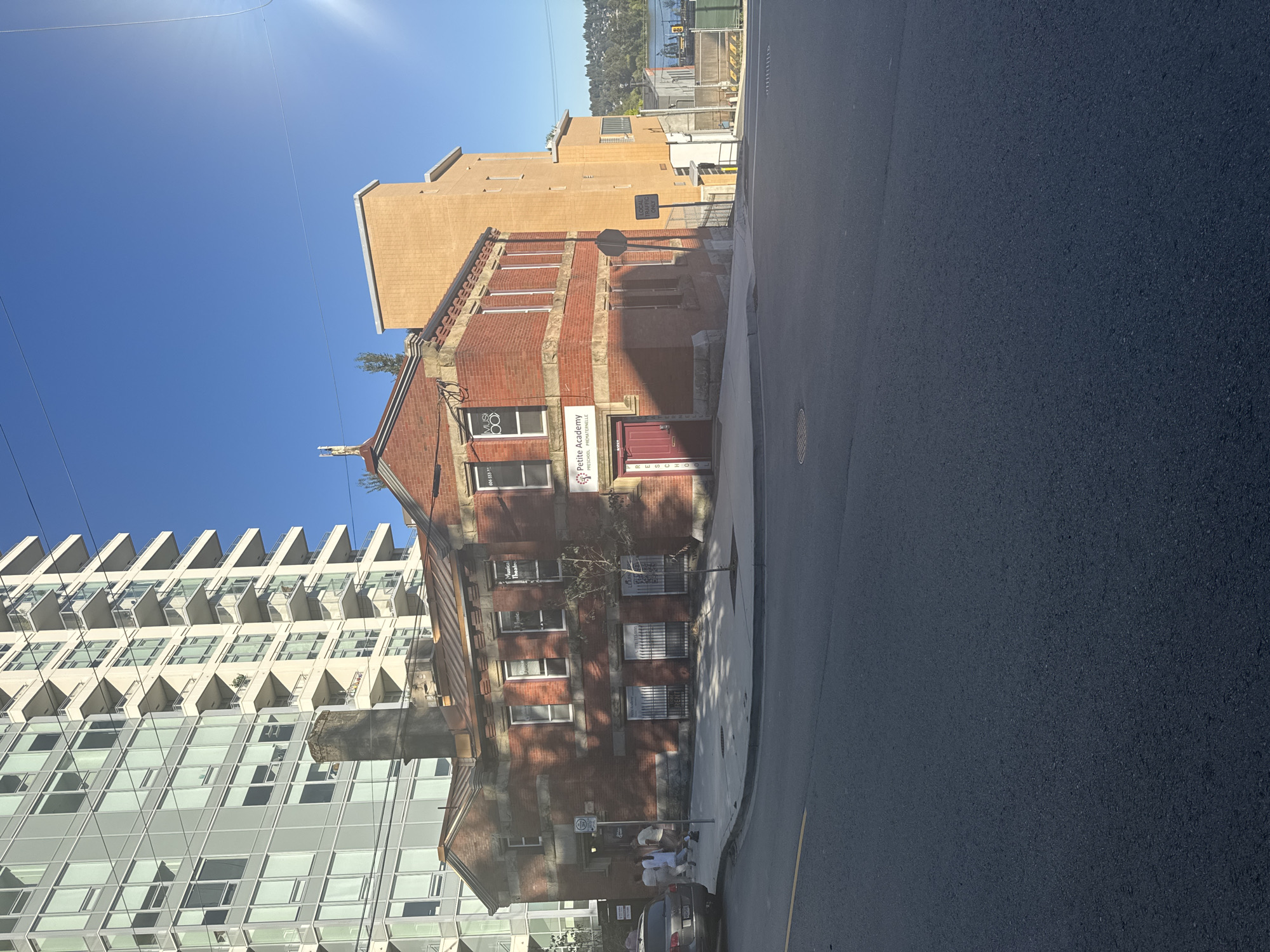 In New Westminster, Canada, an old brick building with a red door and windows advertising "Petite Academy PRESCHOOL PREMATERNELLE" stands on a street corner. A tall, modern apartment building with white balconies rises behind it. Several people are walking near the entrance of the brick building. In the background, a body of water is visible, with trees and hills on the opposite shore. The sky is clear and blue, with the sun casting a strong glare in the upper right corner.