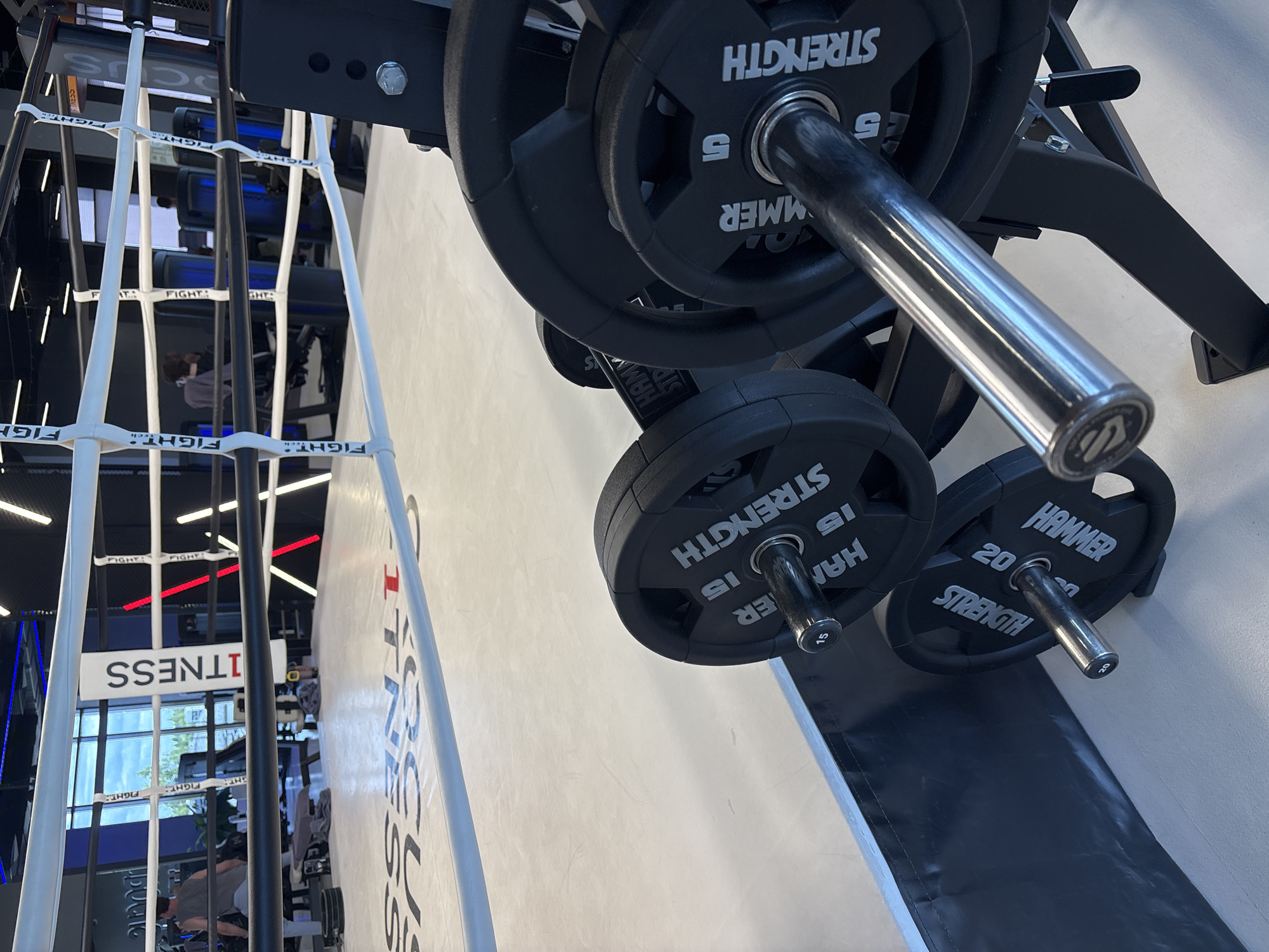 The image shows a gym with a boxing ring in the background and weight plates and a barbell in the foreground. The boxing ring has white ropes and is branded with "FITNESS" and "FIGHT". The floor of the ring has "FOCUS FITNESS" printed on it. In the foreground, several black weight plates are stacked on a rack, with a barbell resting on the rack. The plates have "HAMMER STRENGTH" and their respective weights (15 and 5) printed on them. The gym appears to be modern with bright lighting. There are people in the background, but they are not the primary focus of the image. No specific details about Monolit, Russia are visible in the image.