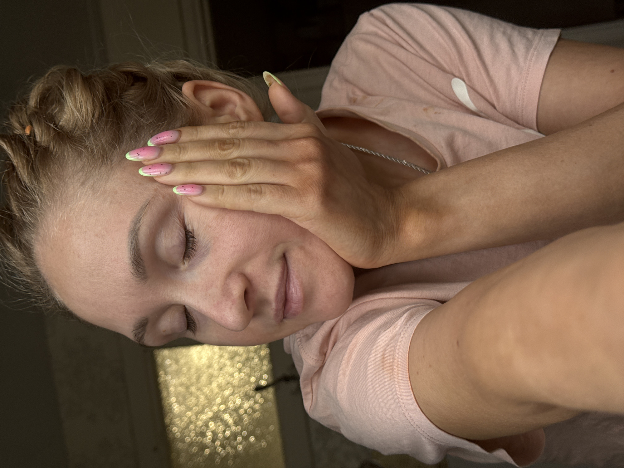 A woman with blonde hair pulled back in braids rests her face on her hand. Her eyes are closed. She is wearing a pink t-shirt and a thin silver necklace. Her fingernails are painted with a pink base and green tips with black dots. The background is out of focus, but appears to show a window with light filtering through. There is no evidence to suggest the location is in Chaikovsky, Russia.