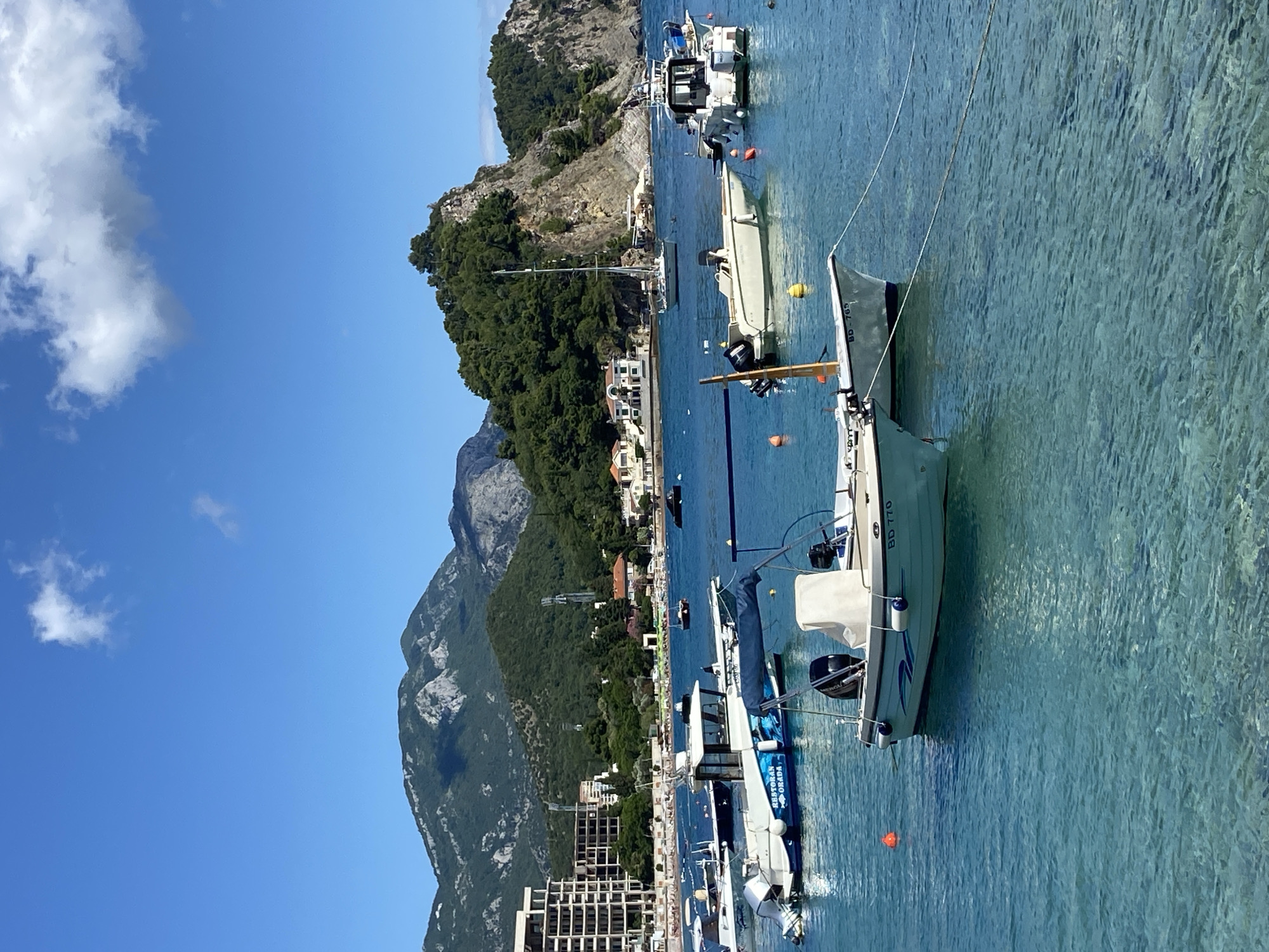 In Petrovac centar, Montenegro, a tranquil bay is dotted with several small boats anchored in clear, turquoise water. The foreshore features a cluster of white buildings nestled against a backdrop of lush green, tree-covered mountains. A modern, multi-story hotel with a white facade is visible on the left side of the image. The sky is a bright, clear blue with a few wispy clouds. The boats, varying in size and design, appear to be leisure crafts, some with outboard motors. One boat in the foreground has a blue stripe and text that reads "RESTORAN GRADA". The clear water allows a view of the seabed in some areas.