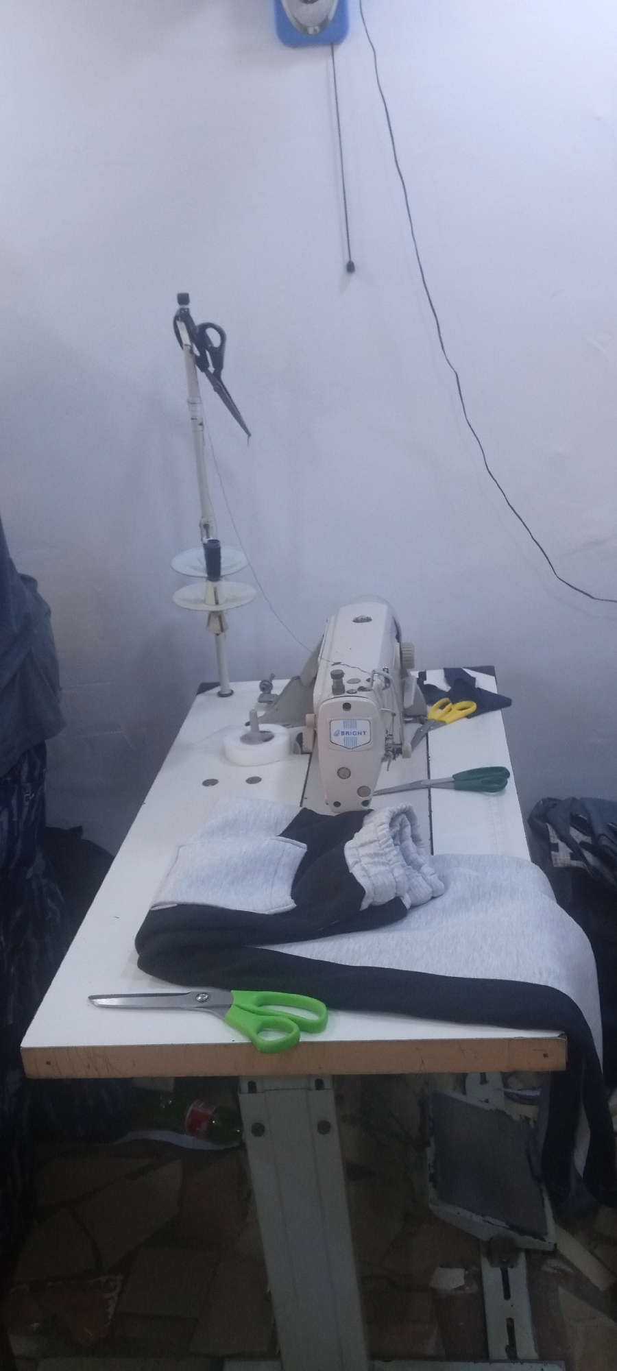 The scene depicts a sewing station, presumably indoors, in Jos, Nigeria.
The primary subject is a white "Bright" brand sewing machine positioned on a white, rectangular table. A spool stand extends upwards from the table behind the machine. Scissors are attached to the spool stand. There is a second pair of scissors on the table beside the clothing.
A partly sewn garment, appears to be a pair of gray and black sweatpants or similar item, sits under the sewing machine.
The table is set against a light-colored wall. A blue steam iron and wire hangs above the sewing station. A green bottle is stored below the table. Piles of unsewn clothing are located on the right and left side of the sewing station. The image has the characteristics of an amateur photograph.