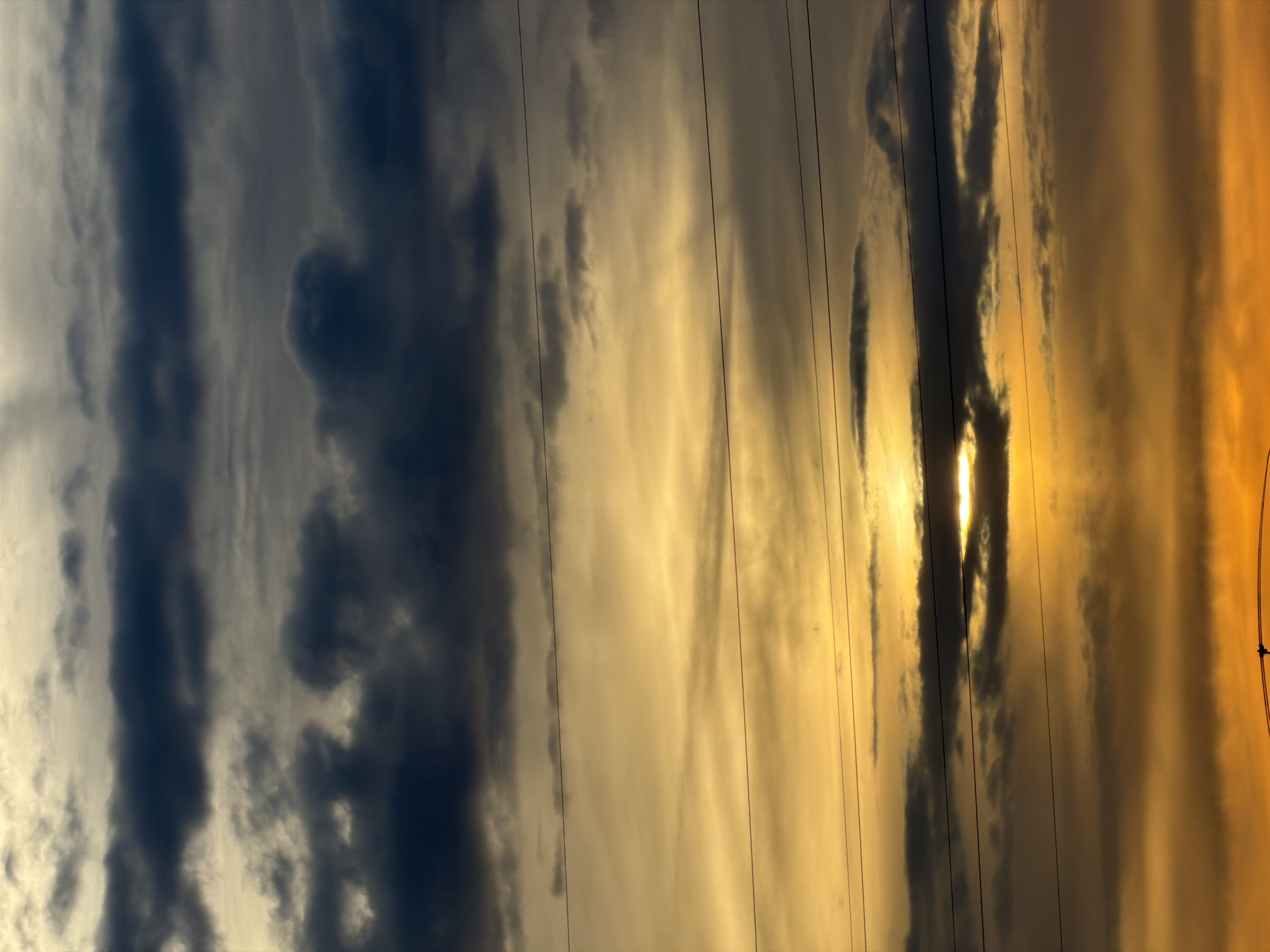 The image depicts a section of the sky at sunset in Esch-sur-Alzette, Luxembourg. The sky has a layered cloud formation, ranging from darker, dense blue-gray clouds on the left to lighter, gold-hued clouds on the right where the sun is setting. The setting sun appears as a bright light source peeking through the clouds, casting a warm glow. A series of power lines run diagonally across the frame. The environment is likely an urban area in Esch-sur-Alzette, given the presence of power lines, but no other structures or individuals are visible.