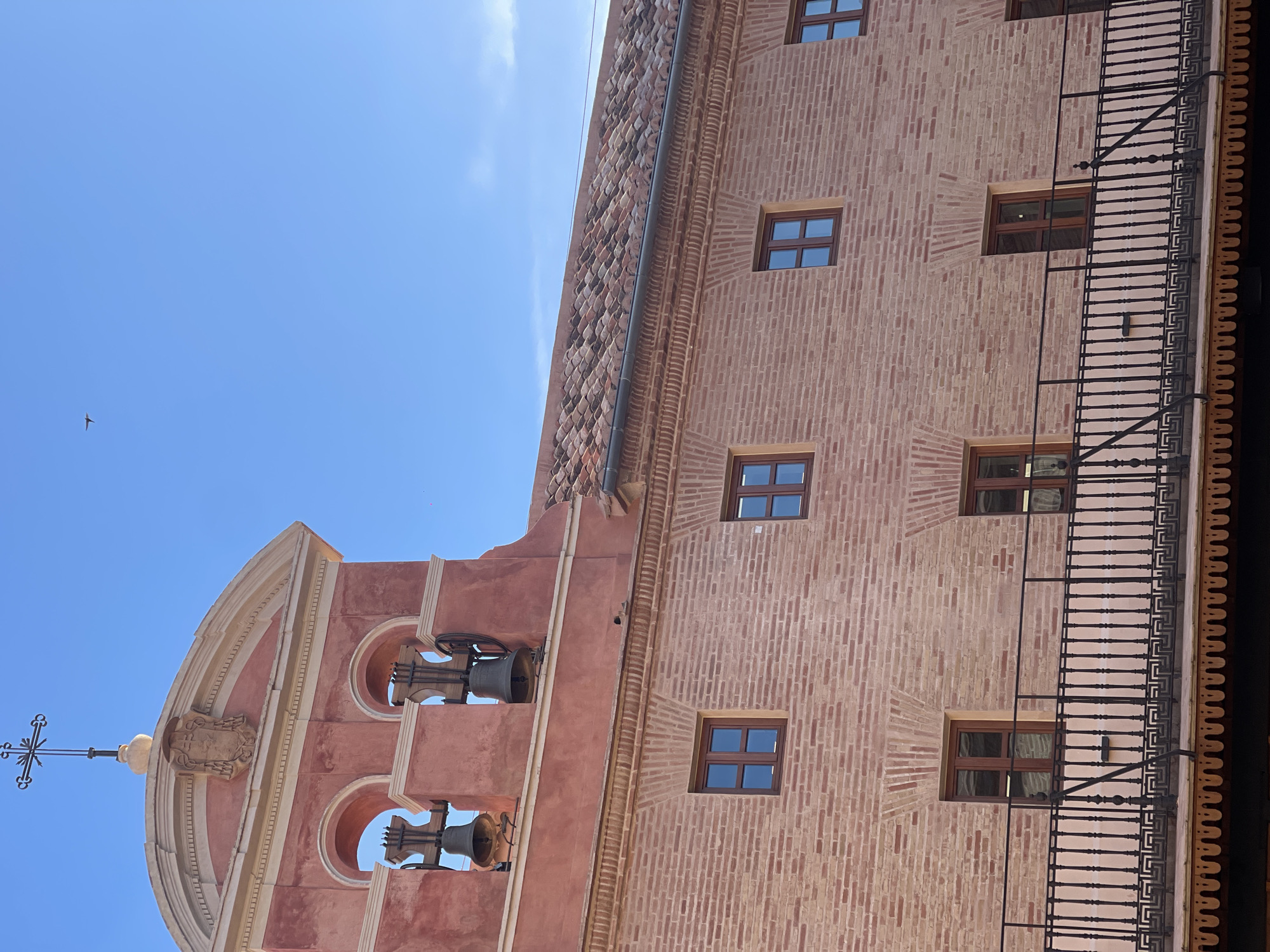 The image shows a section of a building in València, Spain, likely a historical structure. On the left, there is a bell tower featuring two visible bells and an arched opening. The tower is topped with a cross. The main building is constructed of reddish brick, with evenly spaced windows. A black iron railing extends along the side of the building. The sky is clear blue, indicating a sunny day.