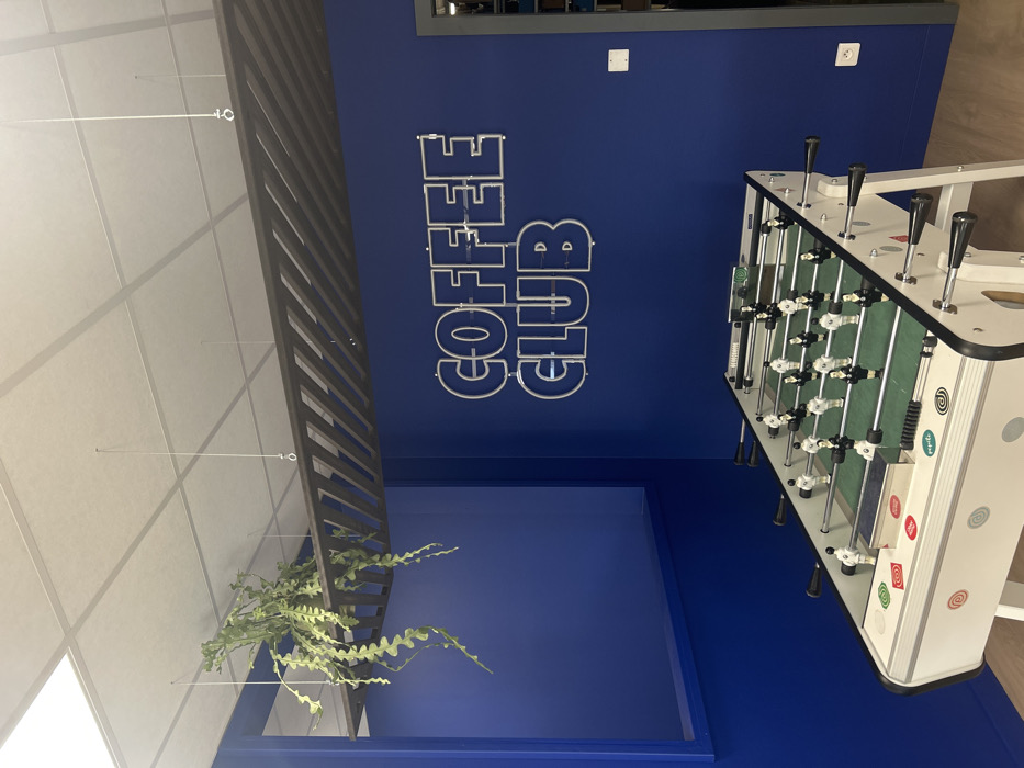 This image depicts an interior recreational space within the city of Reichstett, France. The room features royal blue painted walls and a white grid acoustic tile ceiling. The flooring is a light-toned wood or laminate.
A white foosball table with a green playing surface occupies the foreground and midground, positioned slightly right of center. The table has black and white player figures, black handles, and various colorful circular stickers on its white side panel.
Mounted on the blue wall in the midground are silver-outlined letters spelling "COFFEE CLUB," appearing to be a neon or LED sign. Above this, suspended from the ceiling by thin wires, is a dark-colored, rectangular slatted decorative or acoustic panel. A large, leafy green plant with wavy or serrated foliage, possibly an Epiphyllum anguliger, rests atop this panel on the left side, with its stems cascading downwards.
To the left, a rectangular opening or recess is present in the blue wall. To the far right, a partial view of a doorway with a dark grey frame is visible. A white electrical outlet and a white square light switch are positioned on the blue wall between the foosball table and the doorway. No individuals or active interactions are present in the scene.