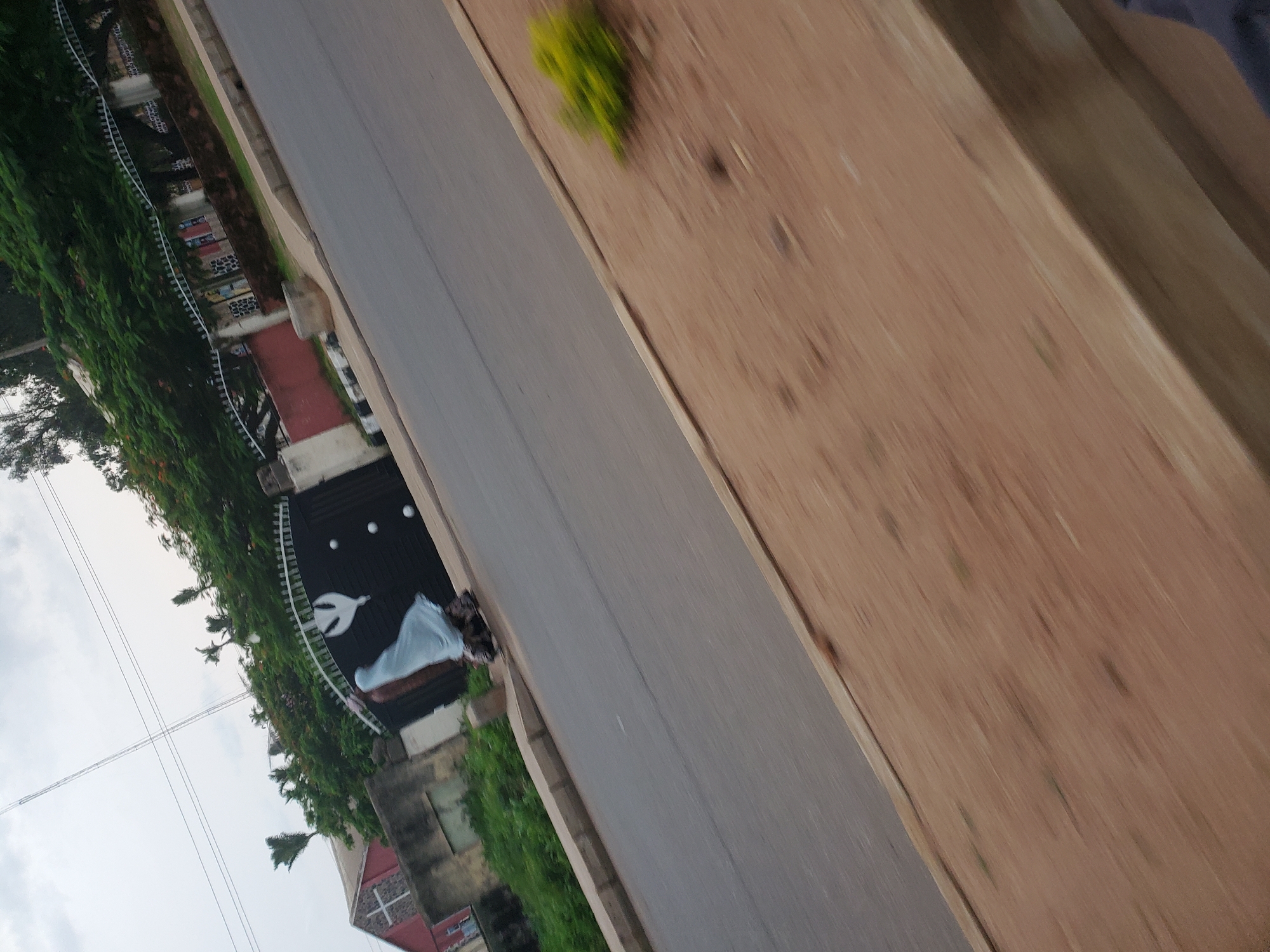 The image, taken in Bukuru, Nigeria, shows a street scene with a black metal gate featuring a white dove emblem. A person wearing a blue veil and a patterned skirt is walking near the gate. A red building with a cross symbol is visible behind the gate, suggesting a church. Trees with green foliage and some orange flowers line the top of the gate. The street is paved, and a dirt embankment with sparse vegetation is on the right side of the road. The sky is overcast with thin clouds. The image appears to be taken from a moving vehicle, as indicated by the motion blur.
