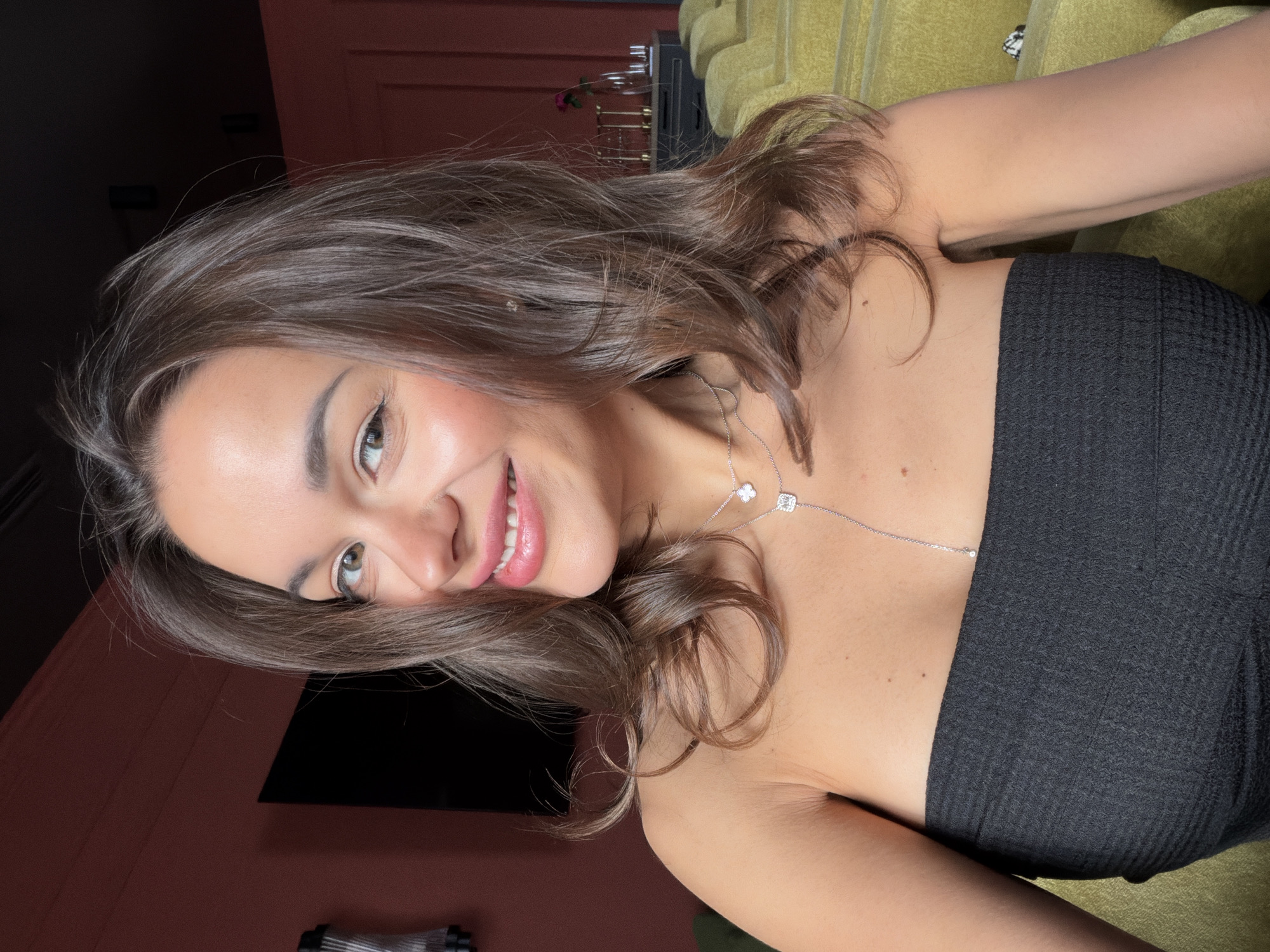 A woman is depicted in a close-up portrait, turned slightly to her left. She has brown hair, light skin, and is smiling. She is wearing a black strapless top and a silver necklace. The background is partially visible and includes what appears to be a green upholstered surface, a dark wall with a television, and a reddish-brown wall. There is no evidence to suggest the location is Krasnoyarsk, Russia.