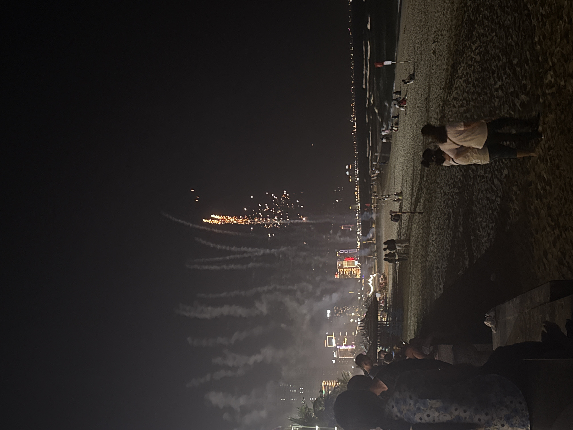 At night in Đà Nẵng, Vietnam, fireworks explode over the city and beach, emitting trails of smoke. People watch the display, many standing on the sand near buildings lining the coastline. Some people are seated on benches or low structures. The beach is illuminated by lights, while the sky above is dark. A couple embraces on the beach.