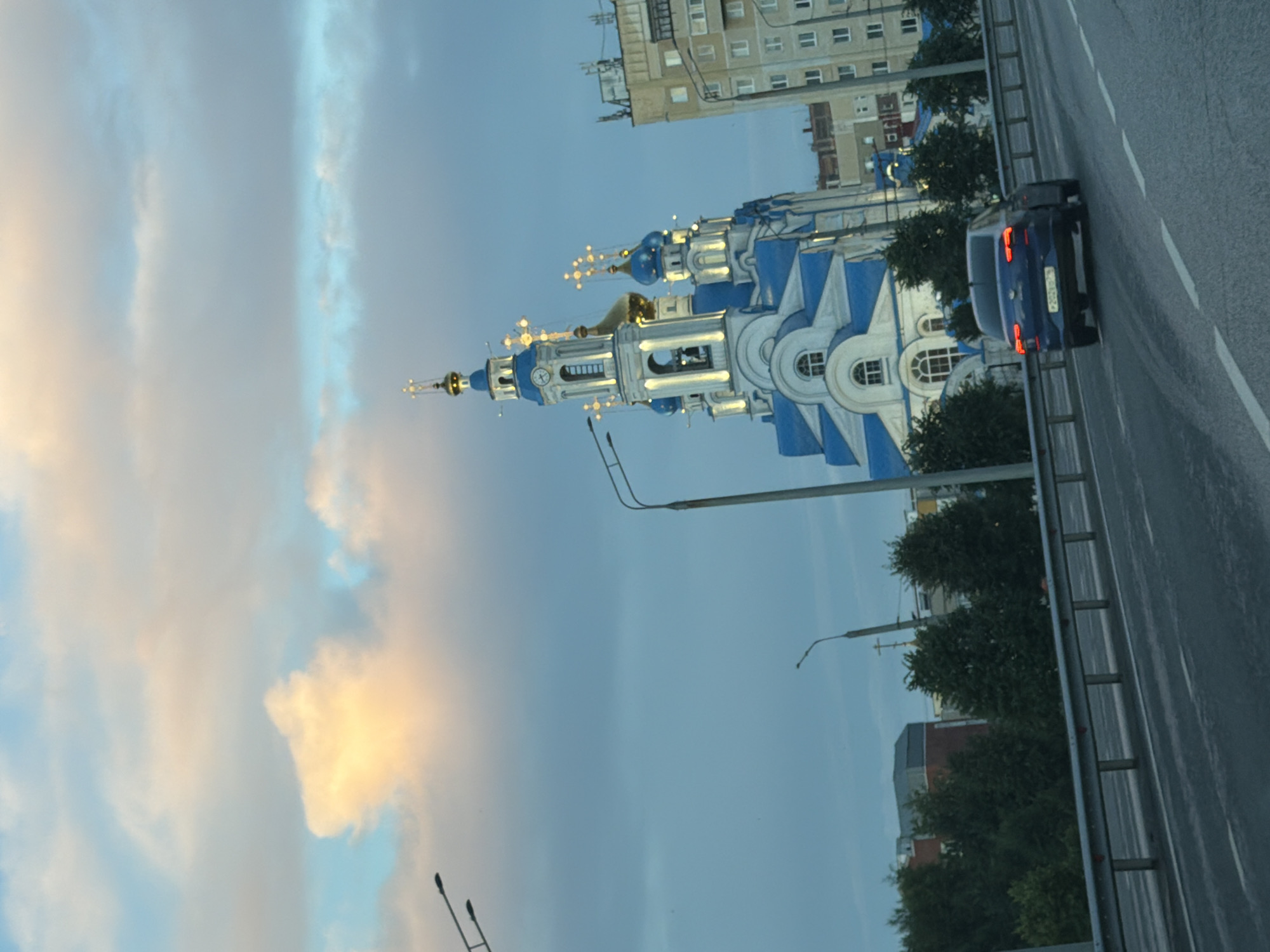The image captures a street scene in what appears to be a city, with a prominent blue and white domed building, likely a church or cathedral, dominating the mid-ground. The building features ornate details, gold accents on its spires, and decorative lights strung across its facade, suggesting it is either decorated for a holiday or illuminated for nighttime. To the right of the building, a dark blue car is driving on a road, its brake lights illuminated. The road is wet, indicating recent rain or a damp surface.
In the background, several apartment buildings with visible windows and balconies are present, typical of urban residential architecture. Trees with dense green foliage are scattered throughout the scene, providing a contrast to the man-made structures. The sky is a soft blue, with wispy clouds, some tinged with a pale yellow from what could be the setting or rising sun, suggesting it is either early morning or late evening. The overall lighting is diffused, contributing to a tranquil or slightly melancholic atmosphere. The image is oriented vertically, with the sky appearing on the left and the road and buildings on the right. No discernible text is visible within the image.