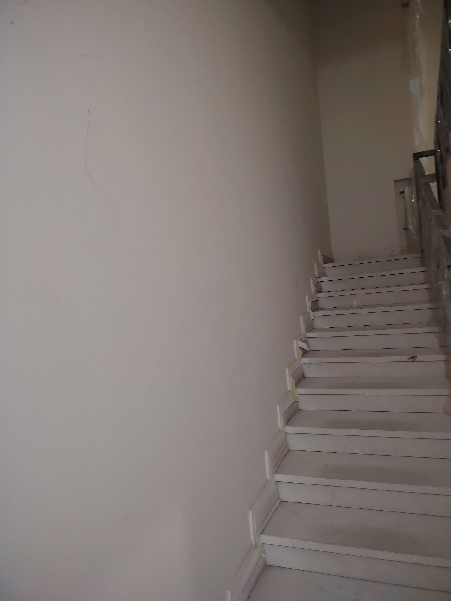 An upward-facing, low-angle interior shot captures a sparse stairwell dominated by a light-colored wall on the left and a functional staircase climbing towards the right. The wall, likely painted off-white or a very light gray, shows several faint scratches and marks, indicating wear or unfinished work.
The staircase consists of multiple steps, possibly made of marble or stone, with risers and treads in a uniform light gray tone. A dark, possibly metallic, handrail runs alongside the stairs on the right, partially obscured and appearing to be wrapped or covered with clear plastic or tape, suggesting either protection during construction or a temporary covering.
The upper section of the image reveals the ceiling line and a corner where the wall meets an upper landing or another wall, which appears slightly darker due to shadows or different lighting conditions. No people or specific activities are visible, presenting a quiet, utilitarian space. The lighting is even, suggesting ambient indoor light, and there is no visible text. The overall impression is one of an unadorned, functional internal passage, possibly in a building under renovation or construction, or a simple, older residential or commercial structure.