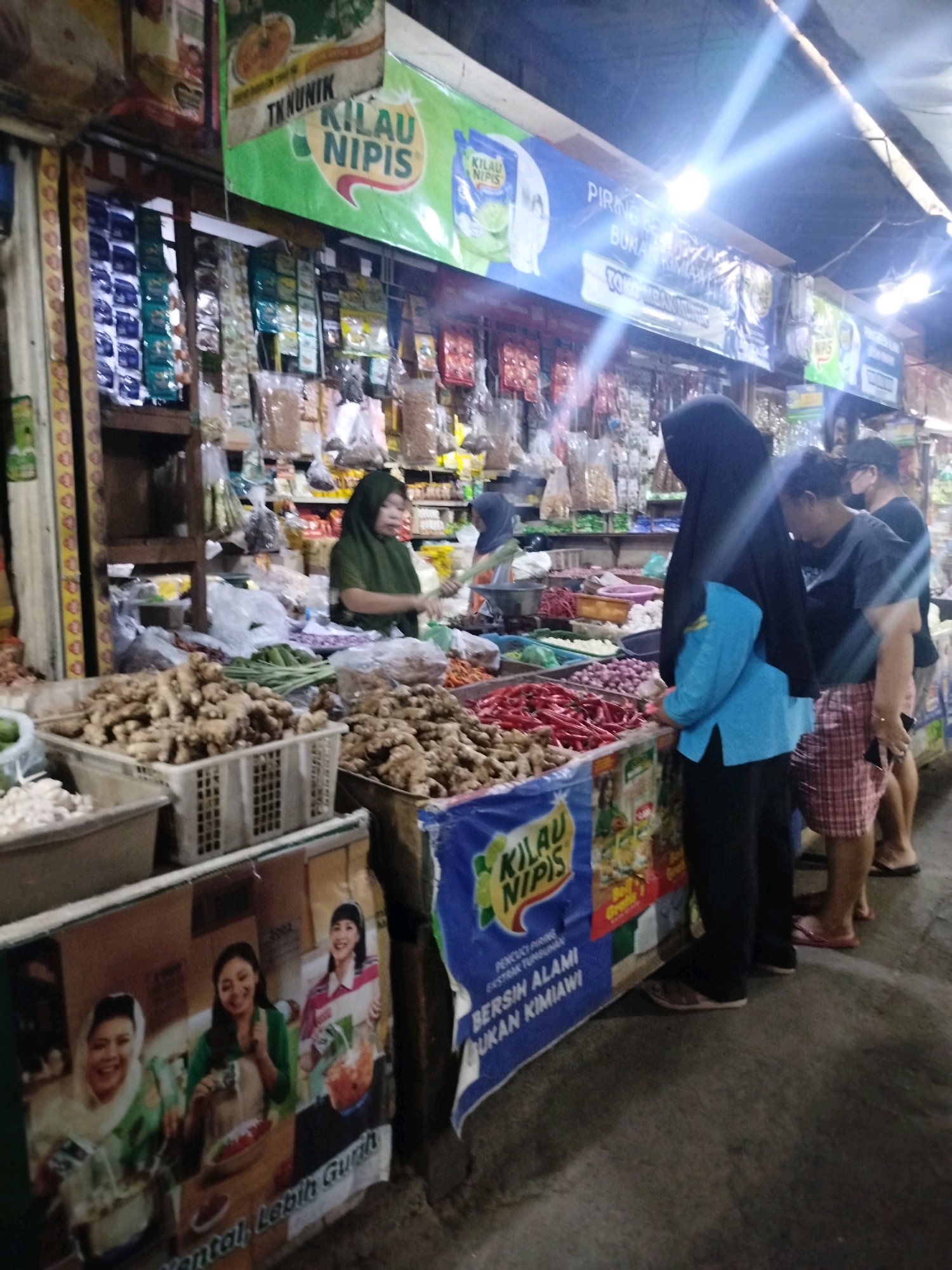 The image depicts a street market scene in Larangan, Indonesia. A vendor, wearing a green hijab and an olive-green top, is seen behind a stall laden with produce. In front of her, baskets overflow with ginger and other root vegetables. To her right, a display of red chilies is visible. Several shoppers are present, including a woman in a black hijab and a blue top, a person in a black t-shirt and plaid shorts, and another person in a black t-shirt. The stall is adorned with various advertisements and banners, one of which features a product called "Kilau Nipis." The environment appears to be an outdoor market with a covered roof, illuminated by artificial lights that cast bright beams. The ground is wet, suggesting recent rain or cleaning. A cardboard advertisement featuring three smiling women is also prominently displayed at the front of the stall. The overall atmosphere is that of a bustling local marketplace.