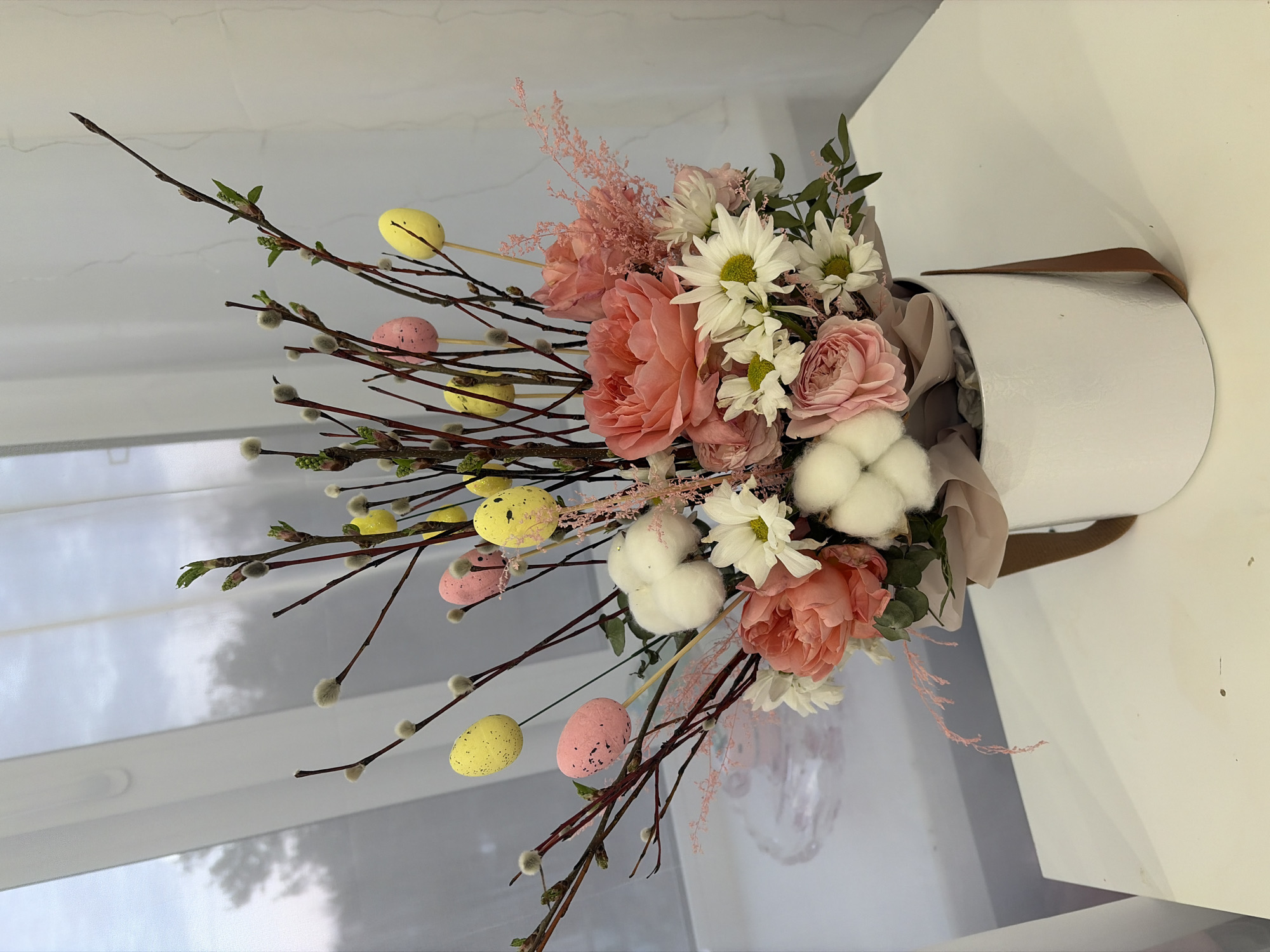The image depicts a floral arrangement in a white cylindrical container. The arrangement includes pink roses, white daisies, cotton flowers, pussy willow branches, and sprigs of pink filler flowers. Yellow and pink decorative eggs are interspersed among the branches. The container appears to be sitting on a white table near a window covered with translucent blinds. There is no indication that the image was taken in Chaikovsky, Russia.