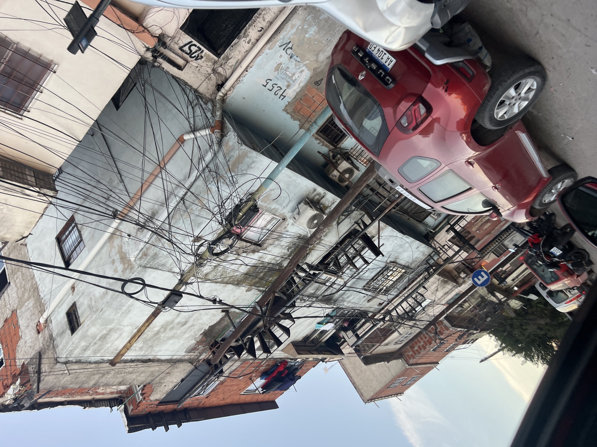 The image depicts a street scene in Buenos Aires, Argentina, characterized by a dense network of overhead power lines obscuring the sky. Buildings of varying heights and architectural styles line the street, many with faded facades and exposed brick. A red Duster SUV is parked alongside the street, its license plate partially visible as "AA 104 FD". Other parked vehicles are also present, though partially obscured. A street sign with the letter "E" is also visible. Laundry hangs from a window, and there is a discernible presence of air conditioning units attached to the buildings. The scene appears to be taken from inside a vehicle, with part of the car's interior visible. The general atmosphere suggests a densely populated urban area with aging infrastructure.