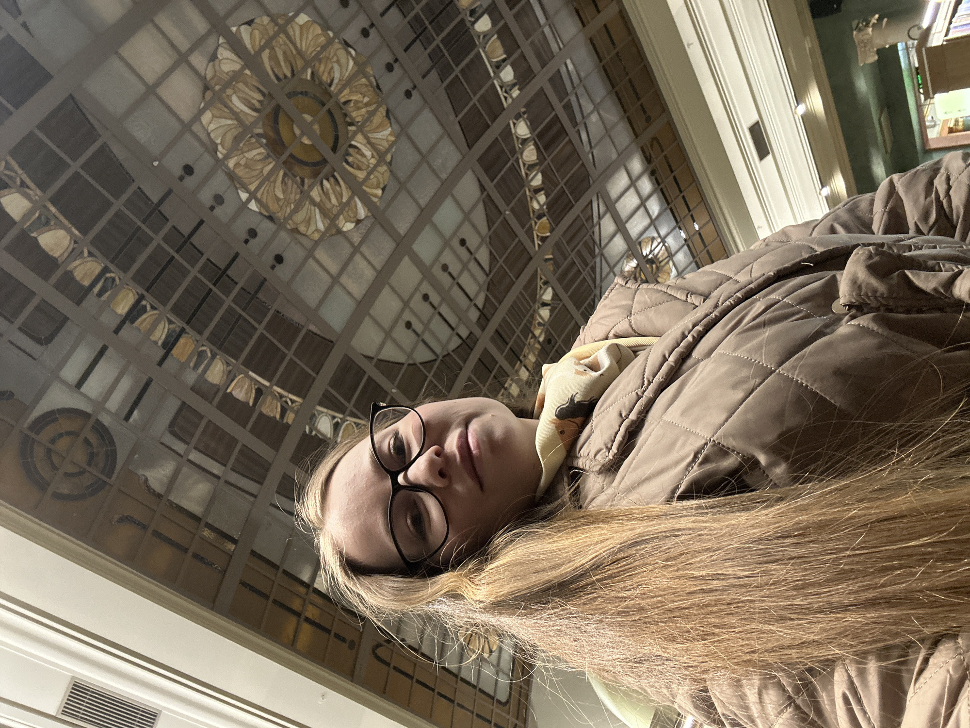 A young woman with long, straight blonde hair and black-rimmed glasses is the central subject, looking directly at the camera with a neutral expression. She is wearing a light brown or taupe quilted jacket and a cream-colored scarf with a subtle pattern around her neck.
The setting is a grand indoor space with a distinctive and ornate ceiling. The ceiling appears to be a large glass or skylight structure, featuring a geometric grid pattern of squares and rectangles. Embedded within this grid are several decorative circular or floral motifs, rendered in shades of gold, cream, and brown, suggesting intricate stained glass or metalwork. One prominent motif is visible to the upper left of the woman, and another partially seen on the right side of the ceiling.
White architectural moldings and trim are visible along the edges of the room, further indicating an elaborate interior. A ventilation grate is also visible towards the lower left. The lighting is soft and ambient, filtering from above through the ceiling, suggesting daytime or a well-lit interior. The overall impression is that of a large public building, such as a historical hall, museum, or grand lobby, consistent with architecture found in Saint Petersburg, Russia. There is no visible text in the image.