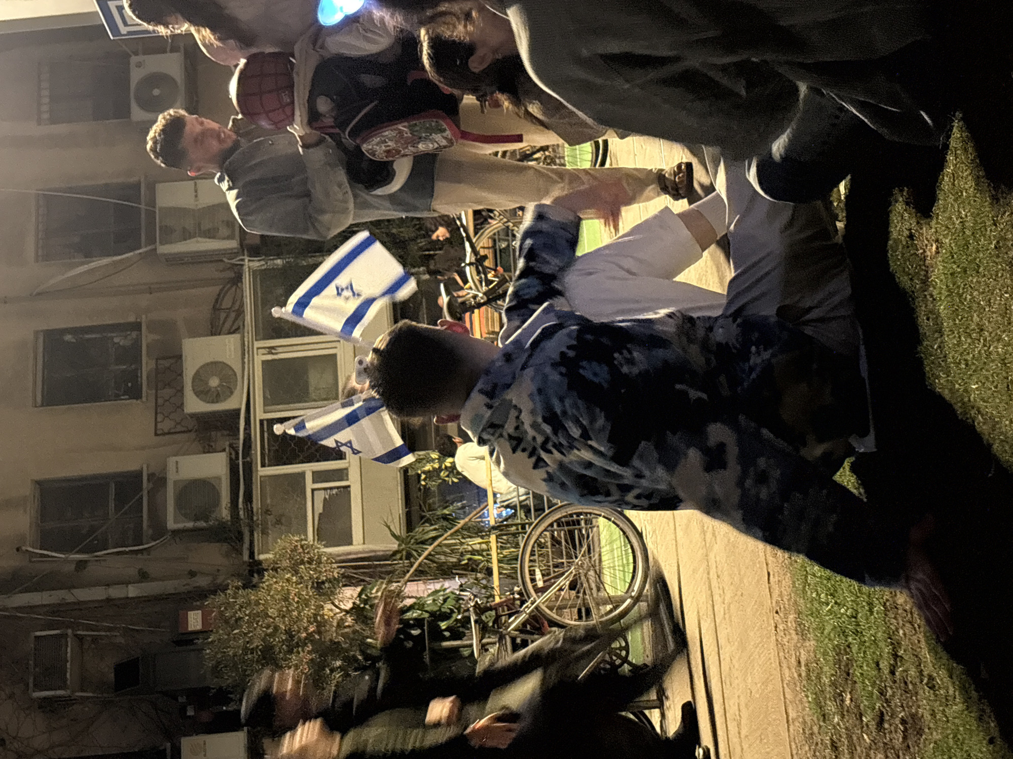 Captured outdoors at night in Tel Aviv, Israel, the image showcases a group of people, some holding Israeli flags, near a building with multiple windows and air conditioning units. A bicycle is also present in the scene.