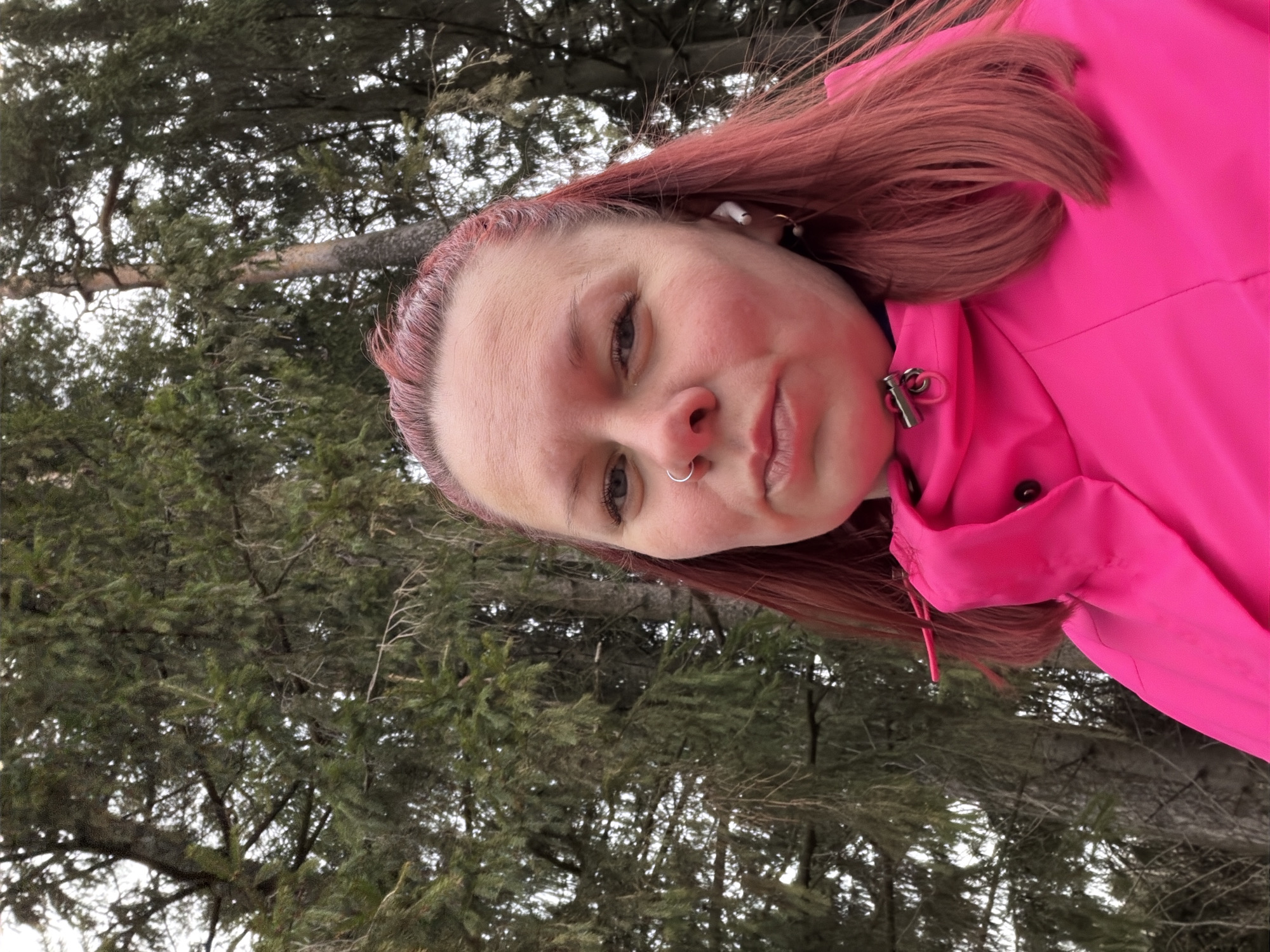 The image is a close-up selfie of a fair-skinned woman with red hair, wearing a bright pink jacket. She has a nose ring and an earbud in her ear. The background shows evergreen trees, suggesting a forest or wooded area. The trees appear to be in Chaikovsky, Russia.