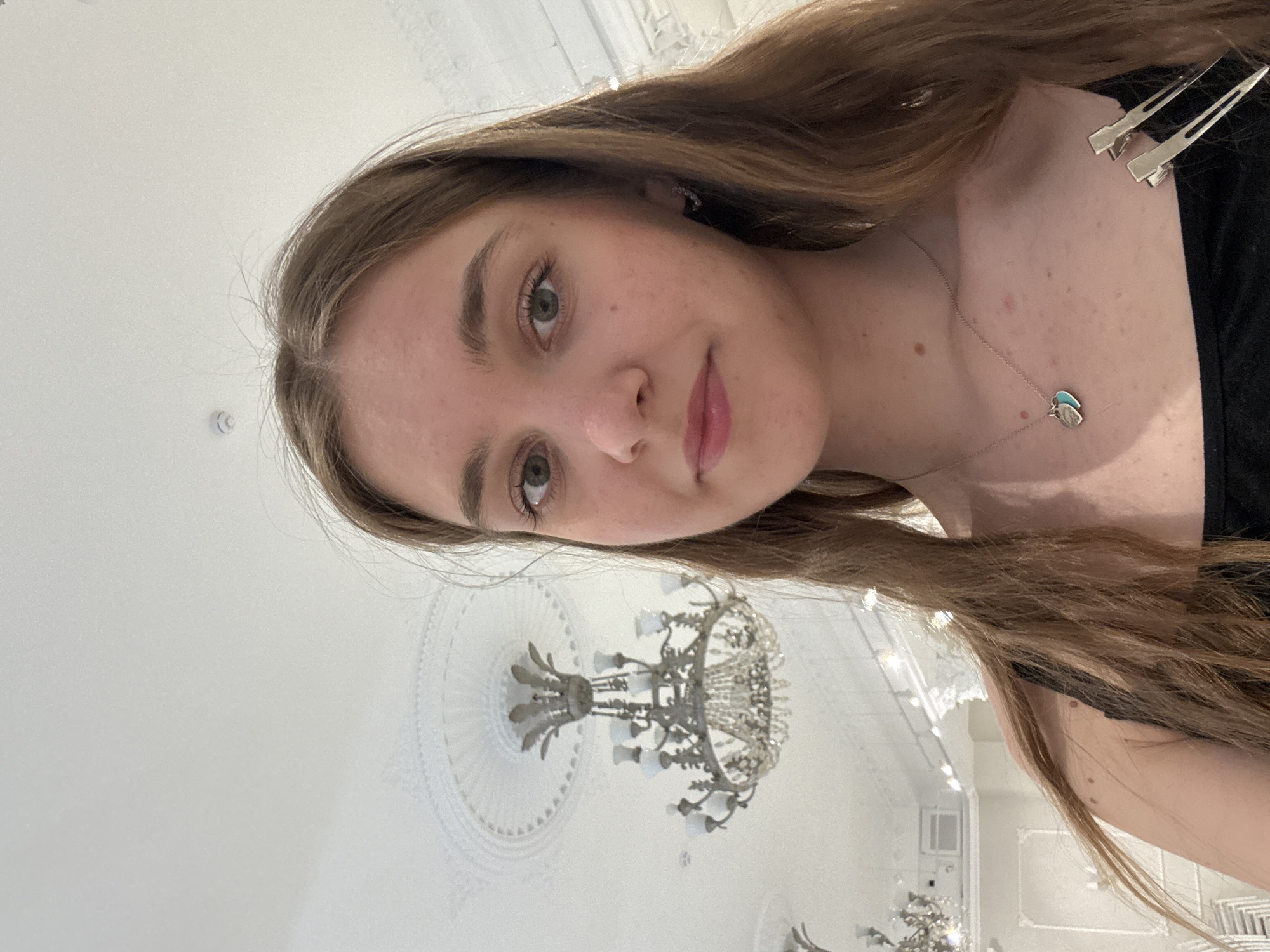 The image is a selfie of a young woman with light brown hair, fair skin, and green eyes. She is wearing a black top and a silver necklace with a blue pendant. Two silver hair clips are visible in her hair on the upper right of the image. The background is a white ceiling with ornate architectural details and a large crystal chandelier. The chandelier, located towards the bottom of the image, is the primary focus of the background. The picture appears to have been taken indoors, possibly in a grand hall or a historic building. While not definitively indicated in the photo, the location is cited as Perm, Russia.