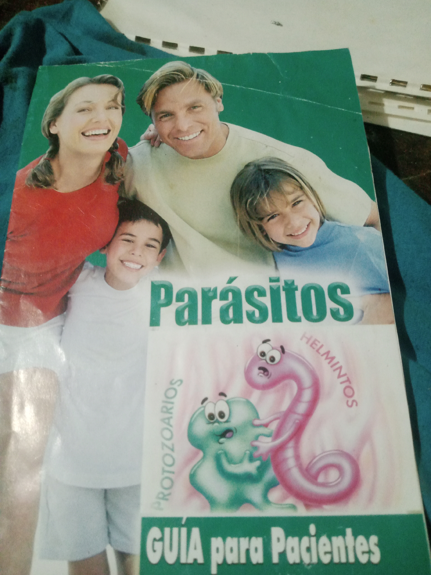 This is a photograph of the cover of a book titled "Parásitos Guía para Pacientes." The cover features an illustration of cartoon-like parasites, one pink and worm-shaped, the other green and blob-shaped, interacting. The text "PARÁSITOS" is prominently displayed in large green letters across the middle of the cover. Below the illustration, the title "GUÍA para Pacientes" is also visible. The background of the cover is a faded green, and the overall image appears to be taken from a slight angle. The photograph itself is of the physical book lying on a dark blue fabric surface, possibly a blanket or clothing. There are no discernible elements in the image that would allow for identification of a specific location within the city of Santa Ana, Venezuela.