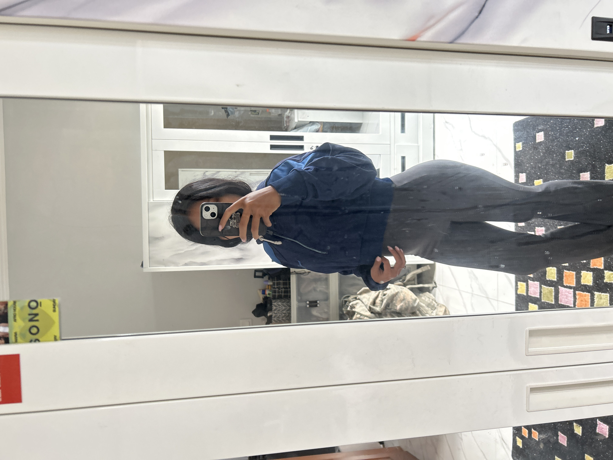 A young woman with shoulder-length dark hair takes a mirror selfie in a room. She is wearing a cropped navy blue jacket and dark grey flared pants. Her left hand is holding a black smartphone with a white case, and her right hand is resting on her hip. Behind her, reflected in the mirror, is a white wardrobe with glass doors, revealing shelves with various items, including baskets and containers. A mat with colorful square patterns is on the floor in front of the wardrobe. To the left of the wardrobe, a red and yellow sticker is visible on the wall. The room appears to be brightly lit. The location is identified as Sukawati, Indonesia.