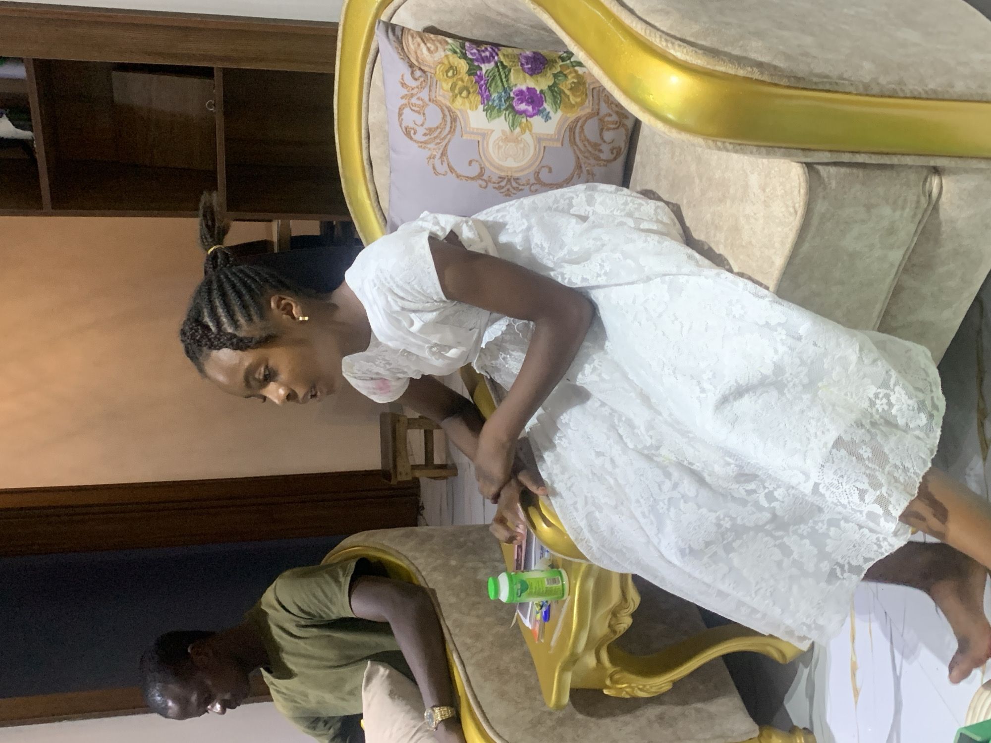 A girl in a white dress sits on an ornate, gold-trimmed chair. A man in a green shirt stands next to the chair. The location is inferred as Jos, Nigeria.