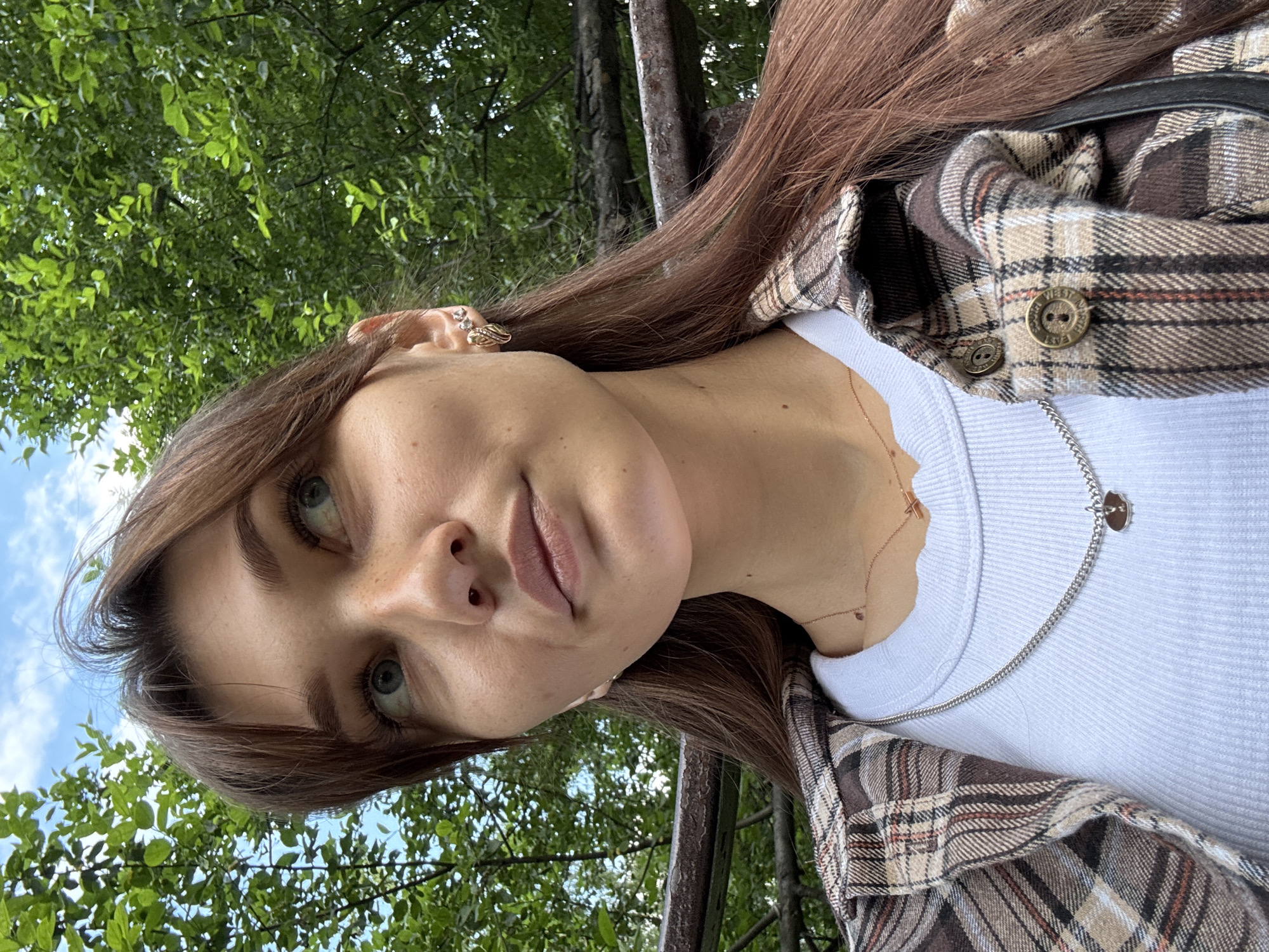 This is a close-up, low-angle selfie of a young woman outdoors, with trees and sky in the background. The woman has long, brown hair, fair skin, and light blue eyes. She is wearing a white ribbed tank top and an open brown and tan plaid shirt. Around her neck, she wears a silver chain necklace with a small, round pendant and another thinner gold chain with a small cross pendant. She has a ring on her left hand and an earring on her left ear. Her expression is neutral, and the lighting suggests it is daytime, likely with bright sunlight filtering through the leaves. The scene appears to be a park or wooded area, with lush green foliage and visible tree trunks. No text is visible in the image. The provided context indicates the location is Novokuznetsk, Russia.