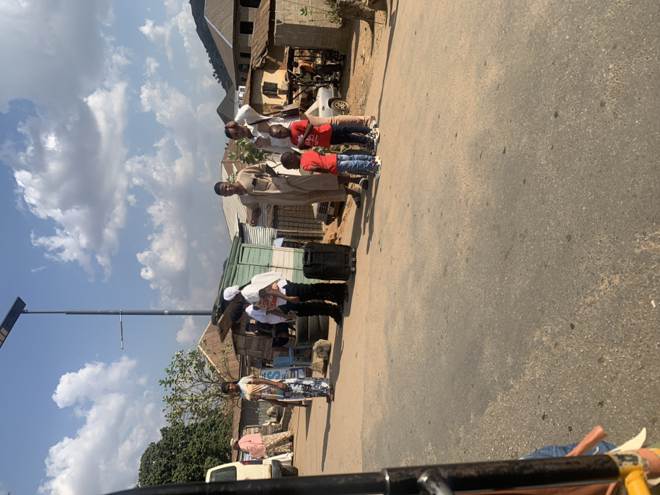 The image depicts a daytime roadside scene in Okengwe, Nigeria. A paved, two-lane road occupies the foreground and midground, with dirt shoulders bordering it. Under a sky with scattered white clouds, a tall, dark grey street light pole equipped with a solar panel and a luminaire stands in the center-left.
On the right side of the road, a group of four individuals is present: an adult male in a light brown tunic gestures with his right hand, standing beside an adult female in a striped top and light-colored trousers who holds her left hand near her forehead, possibly shielding her eyes. In front of these two adults stand two young boys, both wearing red shirts. To their immediate left, a black rolling suitcase rests on the ground.
Further left, another group includes an adult male wearing a white head covering and dark clothing, holding a small child dressed in white. A stack of vehicle tires is visible behind them. To the far left, an adult female wearing a multi-patterned top and trousers stands facing the road. Partially visible behind her is the front section of a white vehicle, possibly a van, and further back, another adult female in a pink top and patterned trousers.
The background features an array of structures, predominantly single-story buildings with corrugated metal roofs and walls, some appearing as simple stalls or workshops. A light-colored sedan is visible behind the group on the right. In the far distance to the right, more substantial buildings with conventional roofs are seen, backed by prominent hills under the clear sky. Shadows indicate bright sunlight from the overhead.