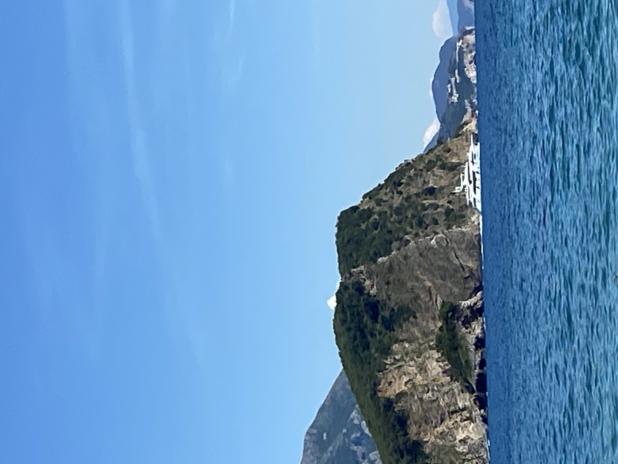 A scenic, medium-shot landscape photograph showcases a section of the coastline near Budva, Montenegro. The image, taken during daylight hours, reveals a combination of rocky terrain, lush vegetation, clear blue sky, and water.
To the left, the image is dominated by a bright blue sky with faint wisps of clouds. A cliff with sparse vegetation, and a mountain range forms the backdrop to the left of the image. The texture of the rocks is visible, adding depth to the scene.
On the right, the sea meets the land. The water is a deep blue, with subtle textures indicating ripples or waves. The coastline is characterized by rocky cliffs, adding a rugged beauty to the landscape.
The overall impression is one of natural beauty, capturing the typical Montenegrin landscape of rugged mountains, a clear sea, and a bright sky. The image is taken with a relatively shallow depth of field, and the colors are vibrant.