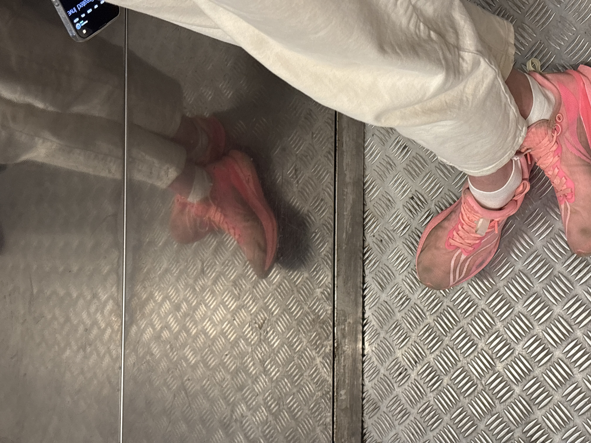 The image depicts a person standing on a metal-plated floor, likely inside an elevator. The person is wearing light-colored, baggy pants and pink sneakers with white socks. Part of a smartphone is visible in the upper left corner. The elevator door and its reflection are to the left, and the floor has a textured, diamond-plate pattern. It is not possible to determine the exact location as being Perm, Russia based on visual information.