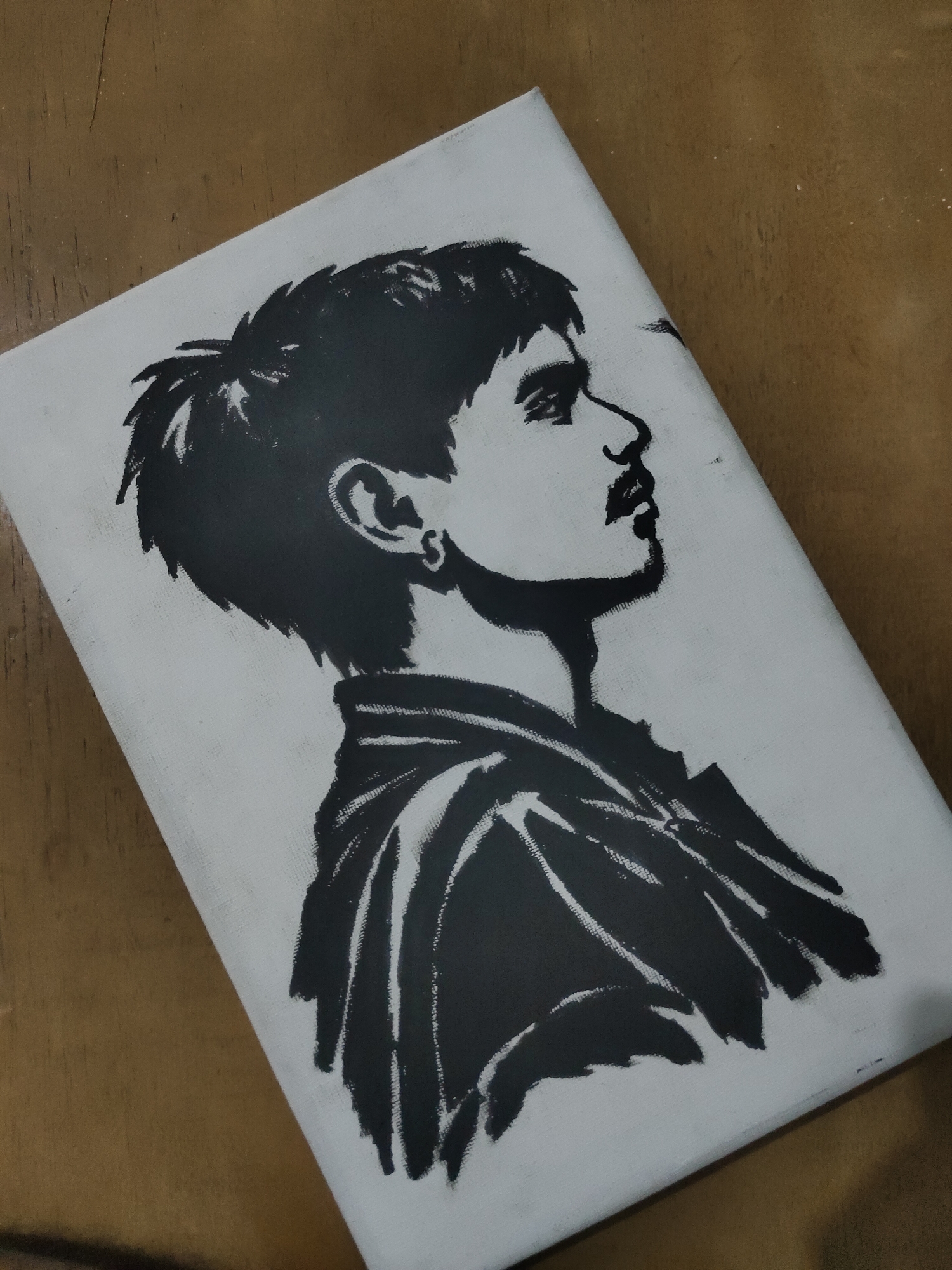 The image displays a white rectangular canvas featuring a high-contrast black and white portrait, resting diagonally on a warm brown, likely wooden, surface. The portrait depicts a young male in profile, facing towards the right. He possesses short, textured dark hair, a prominent nose, full lips, and a distinct dark mustache. A small, round earring is visible in his left earlobe. His attire appears to be a collared garment, rendered with bold, angular black strokes suggesting fabric folds around the neck and shoulder area. The canvas exhibits a visible weave texture. The background surface shows subtle wood grain patterns and minimal scattered white specks. The scene is illuminated by an even, diffused light source.