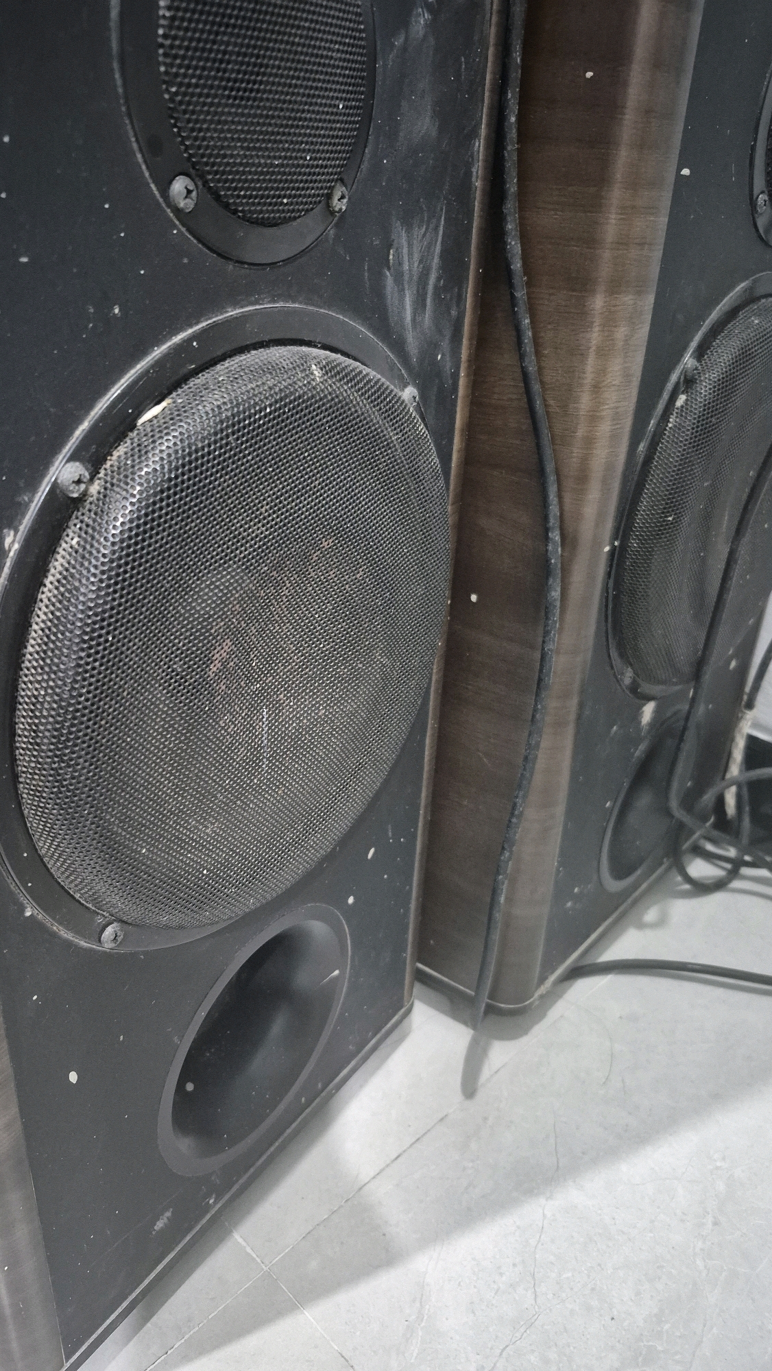 This is a close-up, eye-level, outdoor, daylight, low-angle shot of two dusty black speakers positioned side-by-side on a light gray tiled floor. The speakers have a dark wood-grain veneer on their sides and front panels. The left speaker is larger and shows two circular speaker grilles, one above the other, with a circular port below the lower grille. The top grille is covered by a black mesh and secured by screws. The lower grille is also black mesh, and the speaker cone underneath is visible and appears reddish-brown. The right speaker is partially visible, showing a similar grille and speaker cone. A black cable runs vertically along the seam between the two speakers and extends downwards, with another cable bundled and visible near the base. Dust is visible on the surfaces of the speakers and the floor. The image is cropped tightly on the speakers, with no indication of the surrounding environment in Gambiranom, Indonesia.