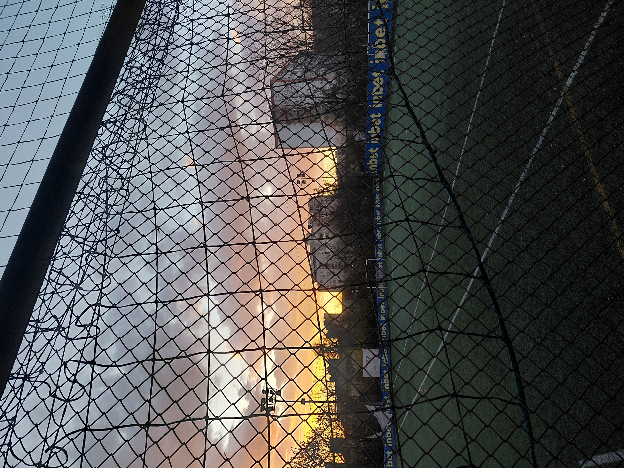 The image, taken through a chain-link fence, presents a scene in Sofia, Bulgaria, at approximately 42.6977° N latitude and 23.3219° E longitude. The view is split, with a sunset or sunrise in the background, illuminating the sky in hues of orange and pink on the left side. The right side reveals an artificial grass surface, likely a sports field.
In the background, through the fence, the outline of buildings and trees are visible. A dark blue panel runs along the fence line on the right side, featuring repeated text "inbet inbet". The fence itself is black, and the mesh pattern obscures the view to varying degrees across the image. The sky to the left of the frame suggests it is either dusk or dawn.