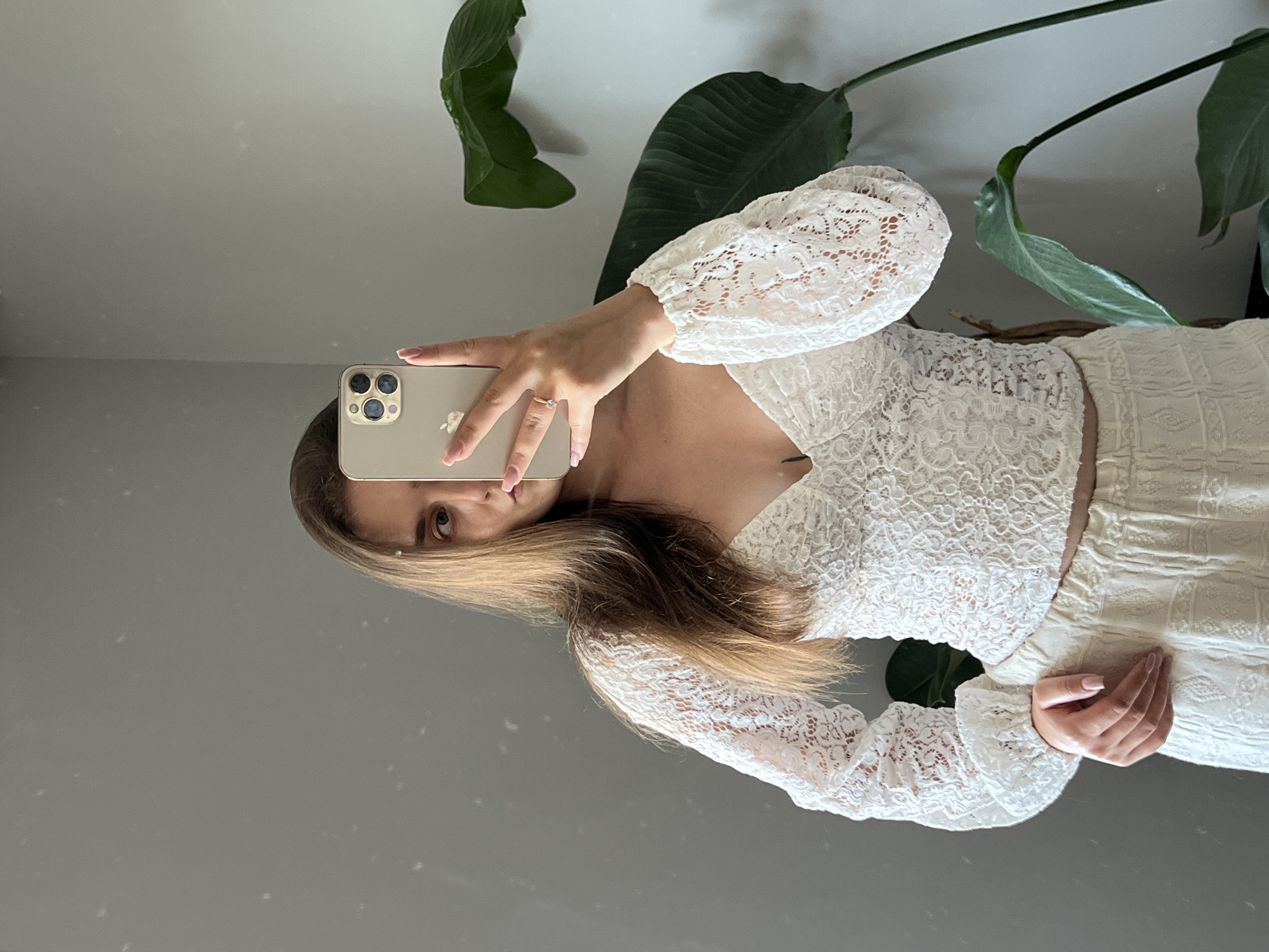 A young woman with long, light brown hair is taking a mirror selfie indoors. She is wearing a white lace top with long sleeves and a V-neckline, and light-colored, textured pants. She is holding a gold smartphone in her right hand, which is angled to capture her reflection. A ring with a small stone is visible on her left ring finger. Behind her, a large, green plant with broad leaves is positioned to her right. The background is a plain, gray wall, which appears to be in a room in Ternopil, Ukraine. There are faint reflections on the wall, suggesting it is a mirror.