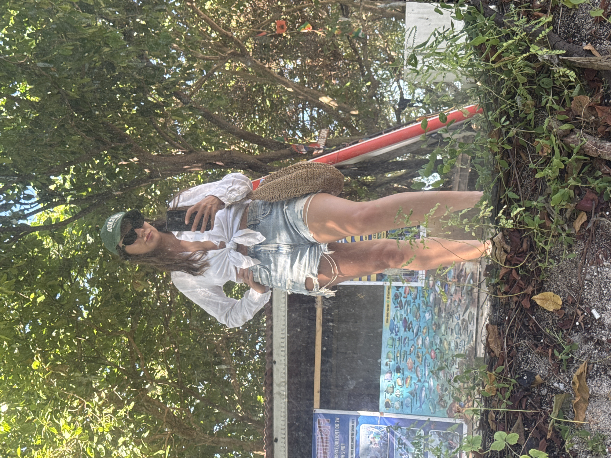 An outdoor mirror selfie captures a woman standing amidst lush greenery, likely in a tropical setting in Indonesia. She is wearing a white long-sleeved shirt tied at the waist, distressed light-wash denim shorts, sunglasses, and a green baseball cap with partially visible text that appears to be "cveo" or "eoo". A woven straw bag is slung over her shoulder. She holds a smartphone up to take the photo, reflecting a sunny daytime scene with bright light filtering through dense tree canopies.
The background features abundant green foliage, including trees and vines, and a natural ground cover of dirt, dried leaves, and small stones. To the woman's right, partially reflected, are two posters illustrating various colorful fish, suggesting a coastal or marine-related activity area. One poster has inverted text, with "FISHING" clearly visible, along with phrases like "WE DO" and "LO'S HO". A red and white object, possibly part of a structure or boat, is also visible in the reflection. The overall atmosphere is relaxed and summery.
