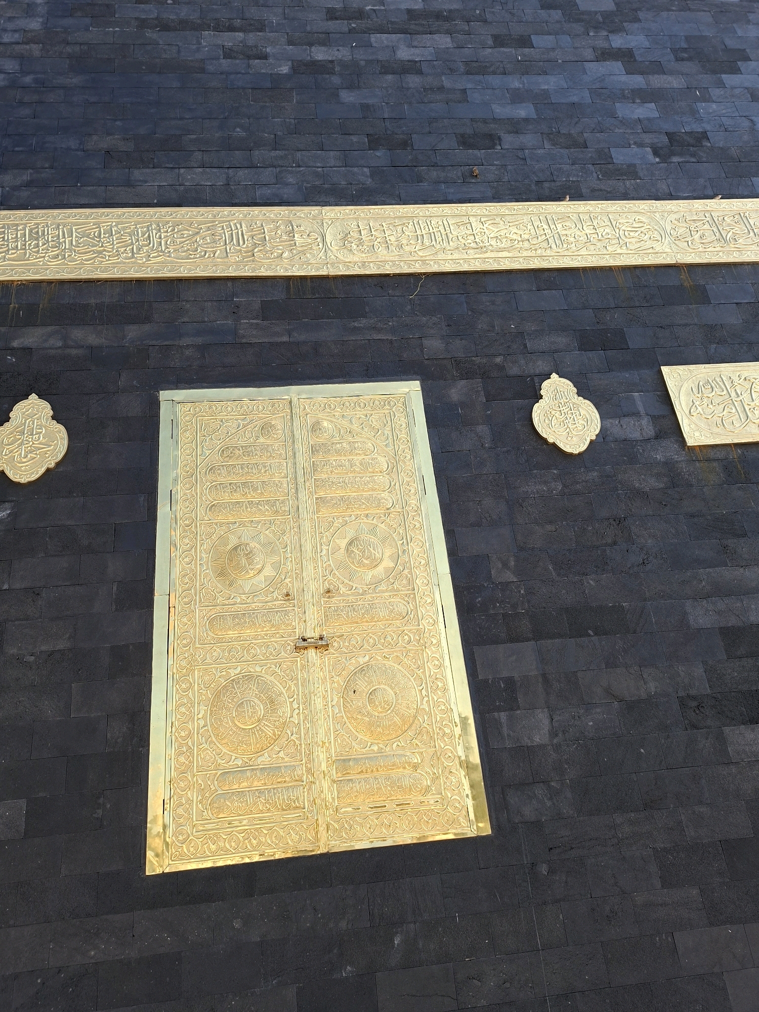 The image displays the exterior of a building with dark, rectangular stone tiles. Prominently featured is a large, ornate golden door, likely a main entrance, with intricate carvings and decorative patterns. Above the door, a wide golden band with Arabic calligraphy runs horizontally across the building's facade. To the left and right of the door, there are smaller, shield-shaped golden emblems also adorned with calligraphy. The lighting suggests it is daytime, with a slight shadow cast on the lower portion of the door, indicating the sun's position. The overall scene depicts a place of significant architectural and possibly religious importance, characterized by its dark stone and opulent golden decorations. This structure is identified as being located in Jrengik, Indonesia.