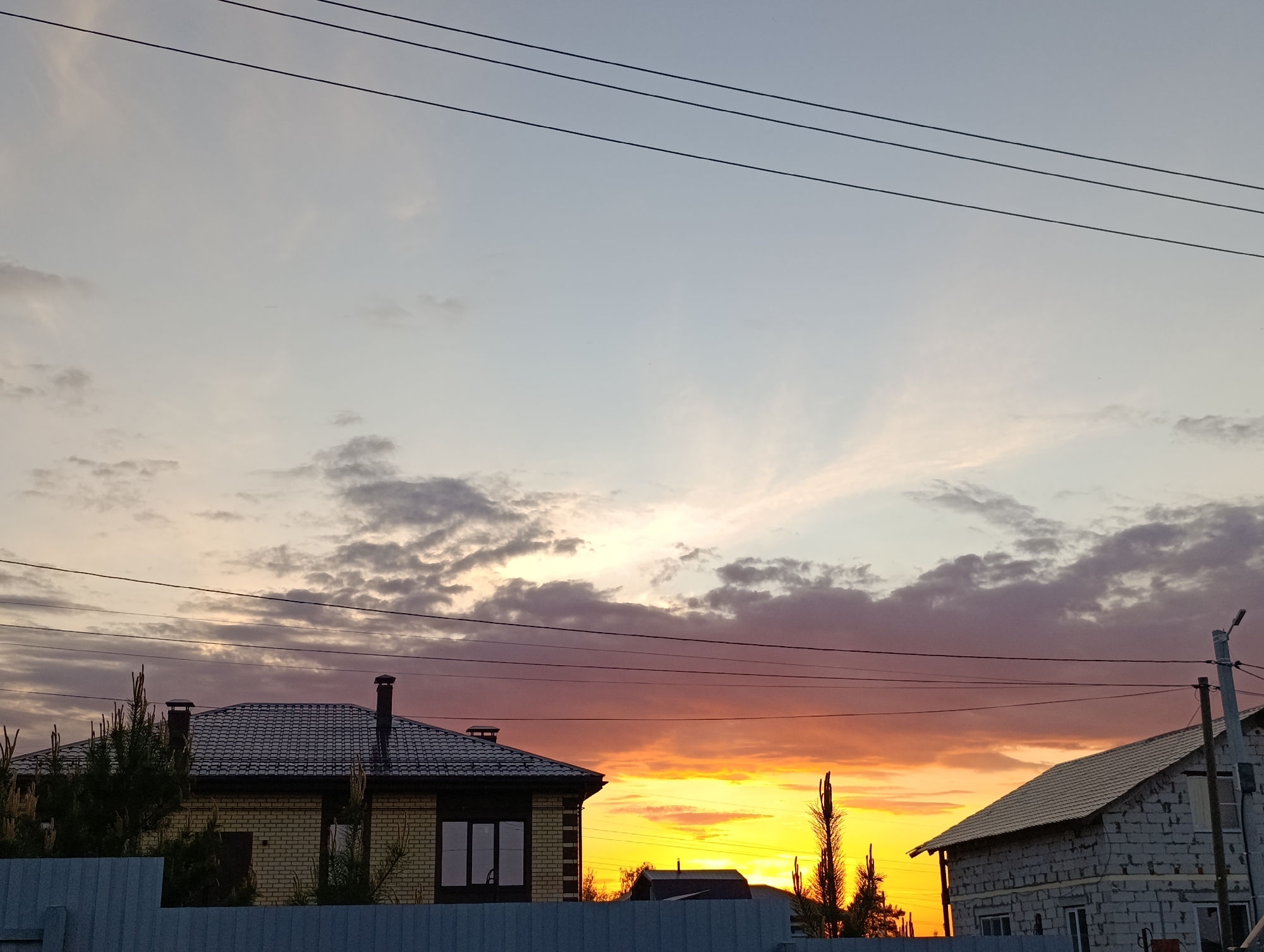 The image depicts a sunset in a residential area, possibly in Chaikovsky, Russia, as per the request. In the foreground, there is a fence, behind which stand two houses. The house on the left is made of light-colored brick with a dark-tiled roof and a chimney. There are some trees or bushes partially obscuring the lower section of the house. To the right is a house constructed from light-gray cinder blocks with a tiled roof. A streetlight is visible next to this house. Overhead power lines stretch across the sky, creating linear elements against the vibrant sky. The sky is the focal point of the image, displaying a vivid sunset with a mix of colors ranging from orange and yellow near the horizon to purple and gray in the upper regions. The clouds are varied, with some dense formations and other thin wisps of cirrus clouds. The sun appears to be setting, casting a golden glow behind the houses.