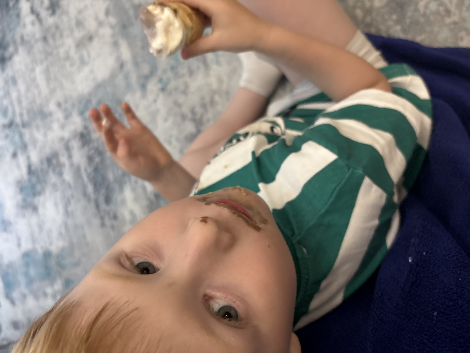 In the provided image, a young child is the central subject, shown from a close-up, upward angle. The child has blonde hair and is wearing a green and white striped t-shirt. Their face is visibly messy with remnants of food, possibly chocolate, around their mouth. One eye is clearly visible, showing a light-colored iris.
The child is holding an ice cream cone in their right hand, which is partially out of focus. Their left hand is also visible, with fingers spread. The background consists of a blue and white patterned surface and a dark blue fabric, likely a blanket or furniture.
The image appears to capture a candid moment, suggesting a relaxed and informal setting, perhaps at home. The food smudges on the child's face imply they are in the process of eating or have recently finished eating. The lighting suggests it might be daytime, but no specific time or weather conditions can be determined. There is no visible text in the image.