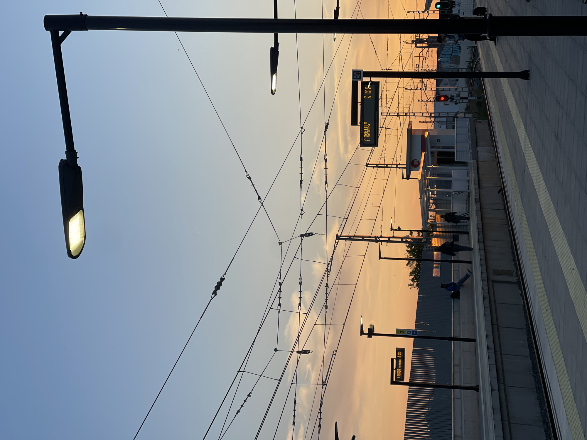 This photograph captures a scene at a train station in Paiporta, Spain during sunset. The sky is gradient, transitioning from blue to orange. Overhead electrical wires crisscross the sky. Several black lamp posts are visible, illuminating the platform. A sign indicates the arrival time of a train to "Maritim" in 7 minutes and "Betera" in 8 minutes. Other people are standing, or sitting on the platform, which is marked with yellow safety lines. In the background, graffiti covers a retaining wall. A red traffic light is visible.