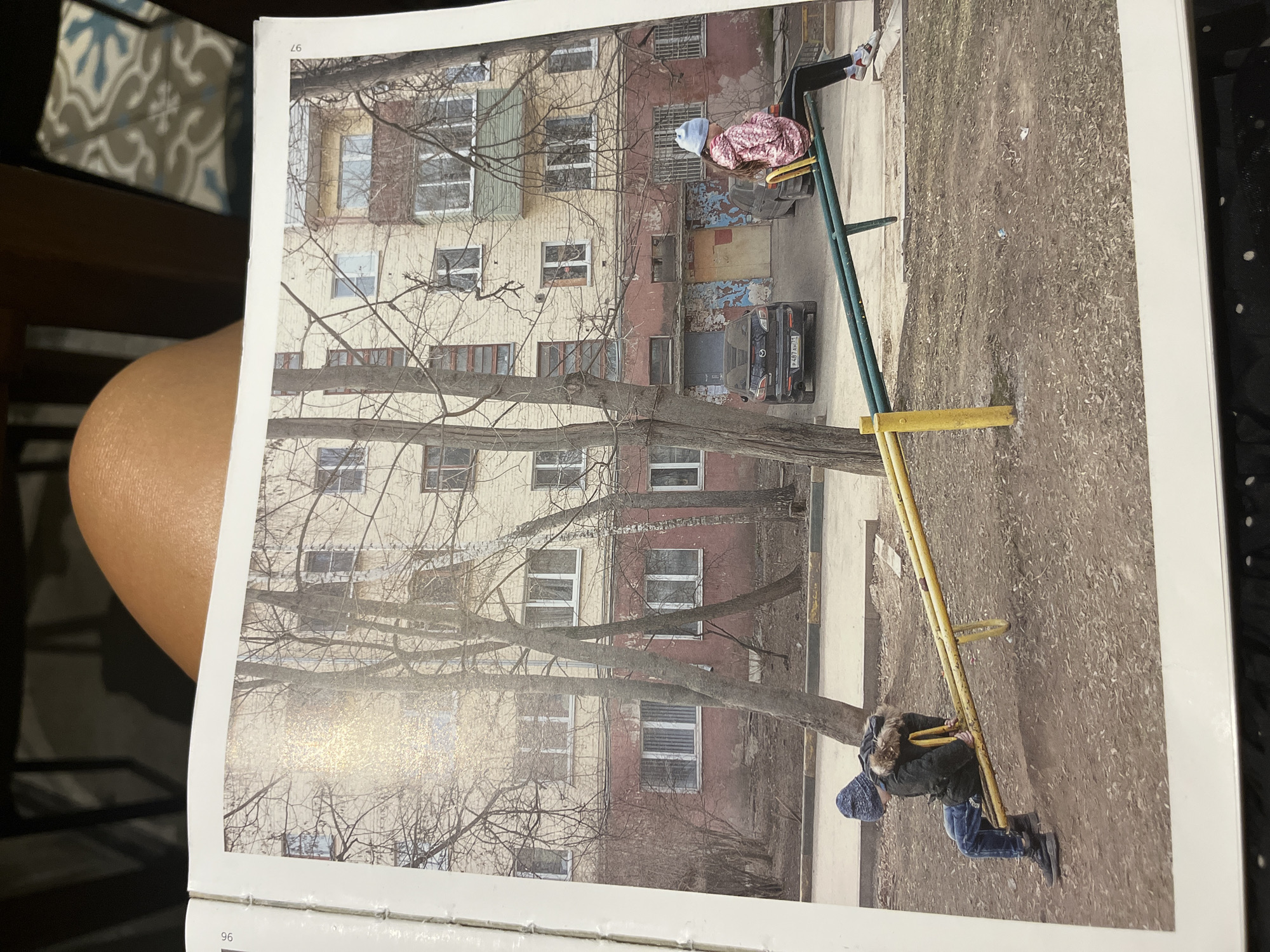 The image shows a photograph from a book of two children on a seesaw in what appears to be an urban environment. The foreground includes the seesaw, which is painted green and yellow, with one child wearing a pink patterned jacket and a light blue hat at the elevated end and another child wearing a dark jacket with a fur-trimmed hood and a blue patterned hat at the lower end. Behind the seesaw, there is a dirt or gravel ground area with scattered debris. In the background is a multi-story building with a mix of beige and red painted sections, with multiple windows. A large tree with bare branches spans across the middle ground. A dark colored car is parked next to the building. The image is a photograph of a page from a book. There is no evidence this is in Ubud District, Indonesia.