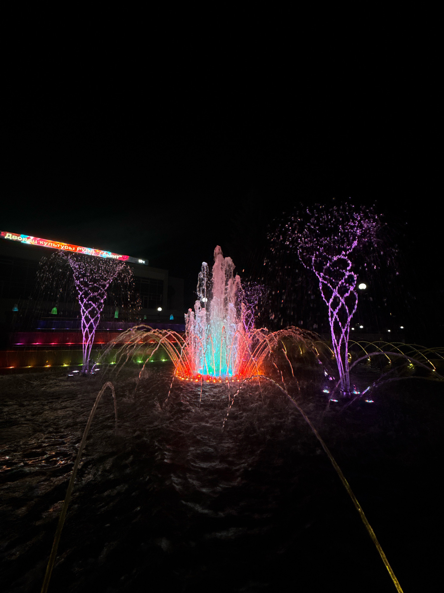 The image depicts a vibrant, illuminated fountain display at night in Mikashevichy, Belarus. The fountain is the central focus, with streams of water shooting upwards and outwards, catching the light from colorful underwater LEDs. The water is illuminated in a spectrum of colors, including bright green, blue, red, and orange, creating a dynamic and mesmerizing effect.
Two stylized, tree-like water jets on either side of the central fountain are lit with purple and pink hues, adding to the artistic composition. These jets resemble shimmering, abstract trees made of water. In the background, a building is visible with a large, illuminated sign that reads "ДВОРЦЫ КУЛЬТУРЫ РУДАК" in colorful, neon-like lettering. The sky is dark, emphasizing the glow of the fountain and the sign. The ground around the fountain appears to be wet and reflective. There are no people visible in the image. The overall scene suggests a public space designed for entertainment and aesthetic appeal, likely a park or a plaza in front of a cultural center.