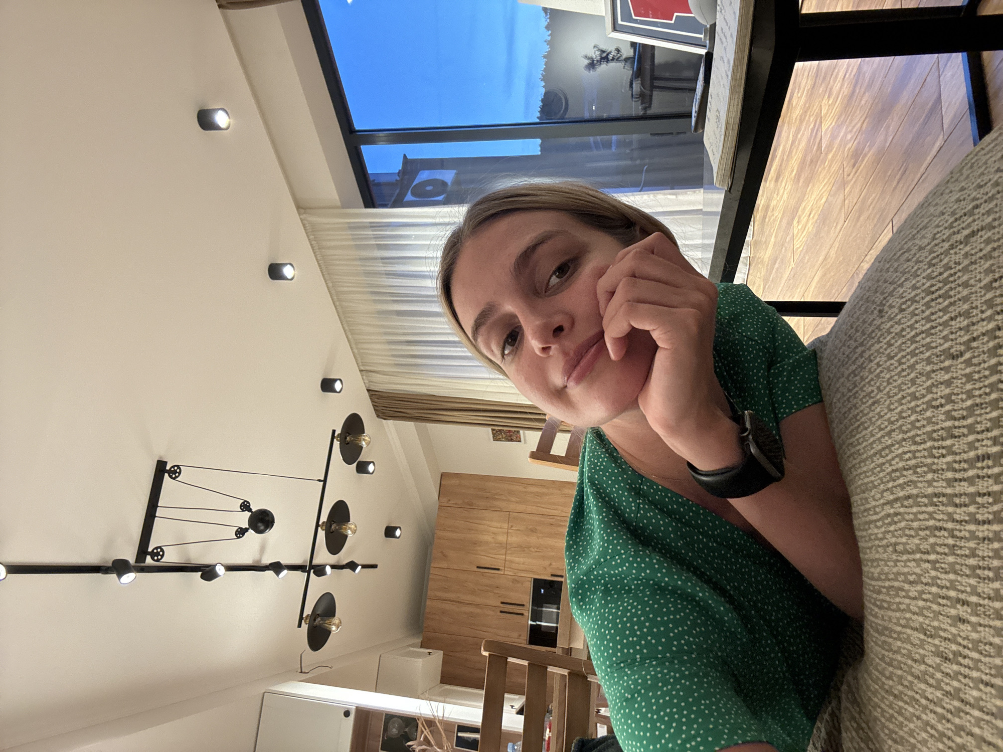 A photograph of a woman with fair skin and blonde hair, wearing a green dress with white polka dots and a black smartwatch, is the central subject. She is seated, resting her chin on her hand, gazing directly at the camera. The background reveals an interior space. Natural light streams in from a window displaying a clear blue sky. The room includes a ceiling with several circular lights, a modern chandelier-style lighting fixture, and a wooden cabinet. A table is visible in the foreground with an upholstered couch. The location is a room with modern interior design, possibly a residential space in an unknown city.