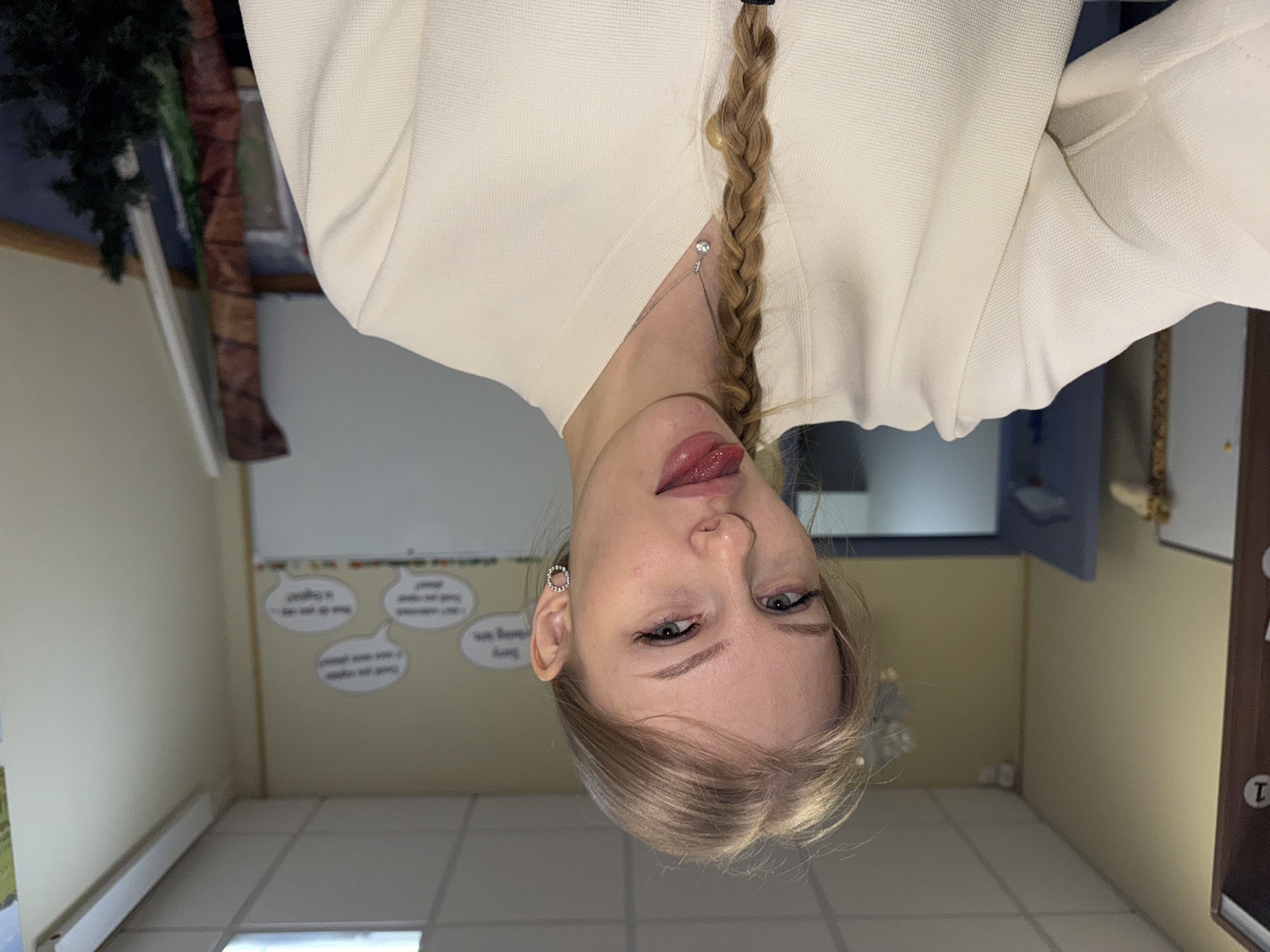 A woman with blonde hair braided down the center is seen from an upside-down perspective, wearing a cream-colored top and a silver necklace. She appears to be indoors, in a room with a white tile floor, light yellow walls, and a white board with speech bubble decorations. A dark-colored door with a circular "1" marking is visible to the right, and a corner with a tree-like decoration is to the left. The location is inferred to be in Perm, Russia.