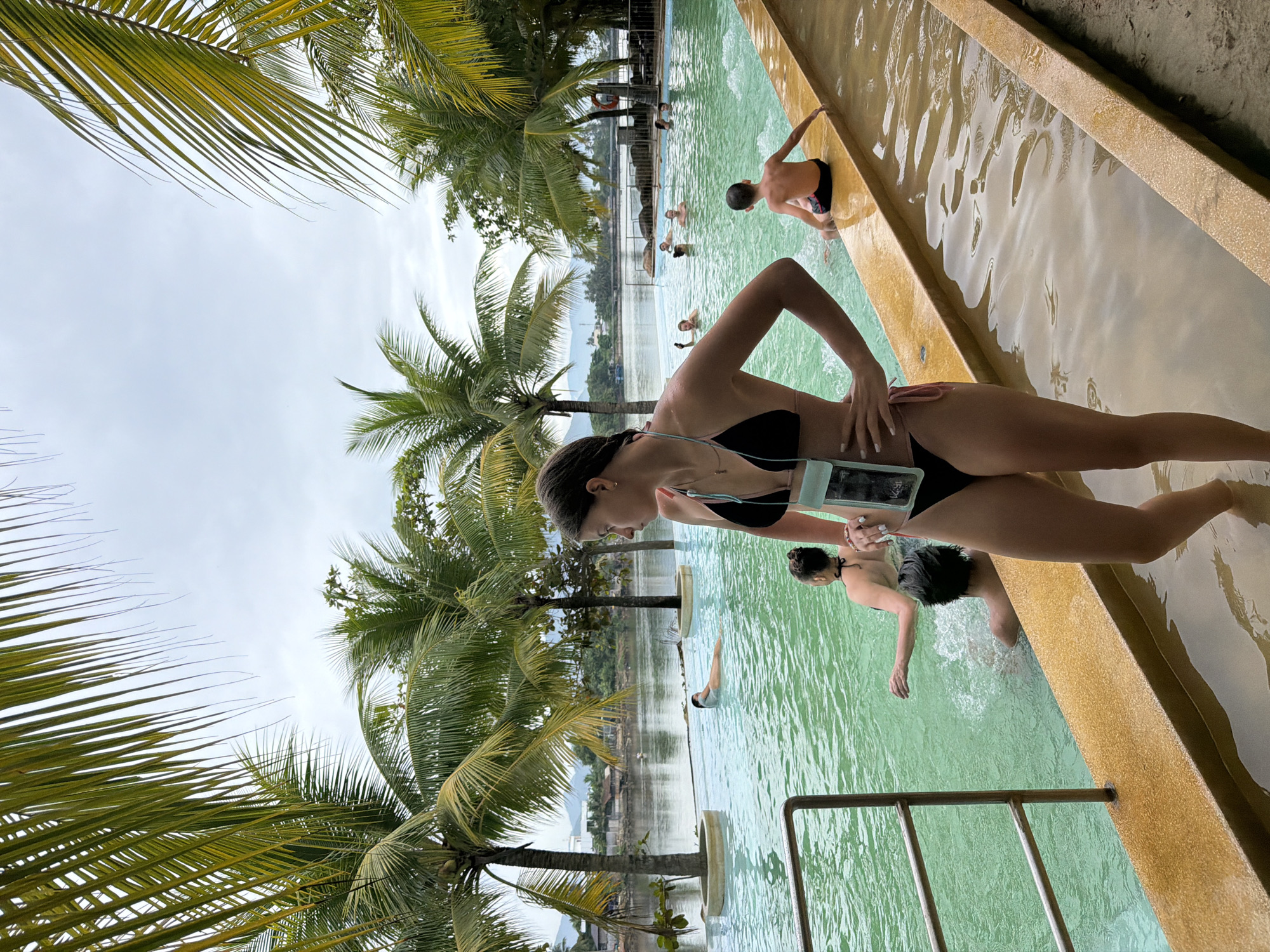 The image depicts an outdoor swimming pool area, likely a resort or a public bathhouse, located in Nha Trang, Vietnam. The pool has clear, green water and is surrounded by a yellow-tiled edge. Palm trees with lush green fronds are abundant, framing the scene and providing shade. In the background, a body of water, possibly a river or the sea, is visible, with distant hills and buildings suggesting a coastal or riverside setting.
In the foreground, a young woman wearing a black bikini stands with her hands on her hips, looking to her left. She has a transparent waterproof pouch with a phone inside hanging from her neck. In the mid-ground, several other people are enjoying the pool. One person is floating on their back, while others are swimming or wading. A young boy is sitting on the edge of the pool in the right mid-ground. The sky is overcast, suggesting a cloudy day. The overall impression is one of a relaxed, tropical vacation spot.