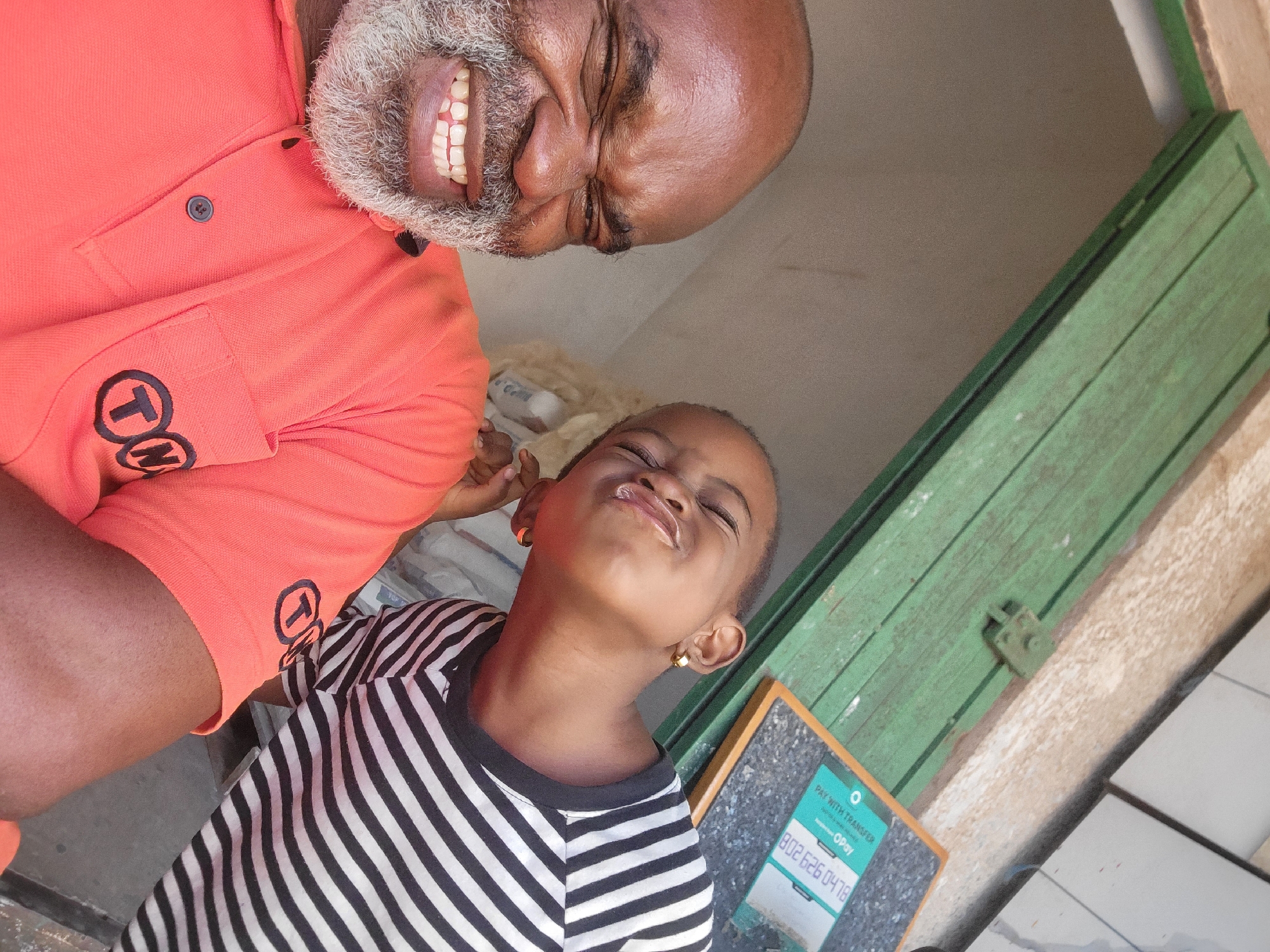 An angled medium shot of a man and a child. The man is positioned in the upper left corner, while the child is in the center-left. The man is older, with dark skin, a grey beard, and a bald head. He wears an orange polo shirt with a logo on the left side. His mouth is open, revealing his teeth. The child, also with dark skin, wears a black and white striped shirt and gold hoop earrings. The child's face is scrunched.
In the background is a white interior with a green wooden door. A sign that reads "PAY WITH TRANSFER" is affixed to the wall below the green door.
This photo may have been taken in Maiburiji, Nigeria.