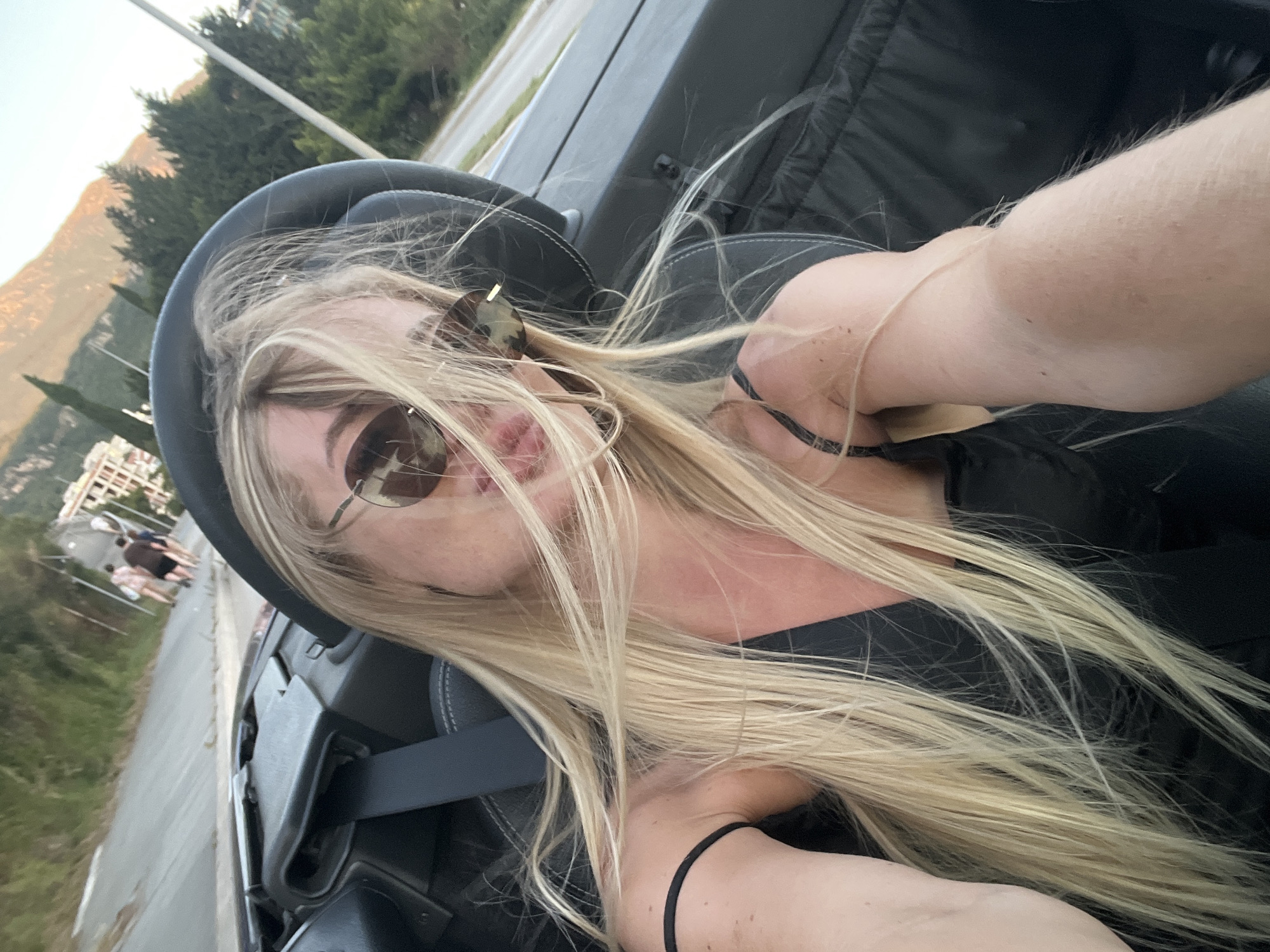 A blonde individual is seated in the driver's seat of a dark-colored convertible vehicle, taking a selfie. Their long, straight blonde hair is windswept, partially obscuring a pair of round, aviator-style sunglasses and a portion of their pink-lipped mouth. They wear a dark, thin-strapped top and a black seatbelt across their chest. A black elastic band is on their left wrist, which is extended, holding the camera. The vehicle's interior is dark, with visible black leather seats and dashboard elements. The scene is set on a paved road in Boreti, Montenegro. In the mid-ground, several indistinct pedestrians walk on a road shoulder, alongside buildings with light facades and red roofs. The far background features large, rugged mountains illuminated by warm, late-day sunlight under a light blue and orange sky, with scattered green trees and utility poles.