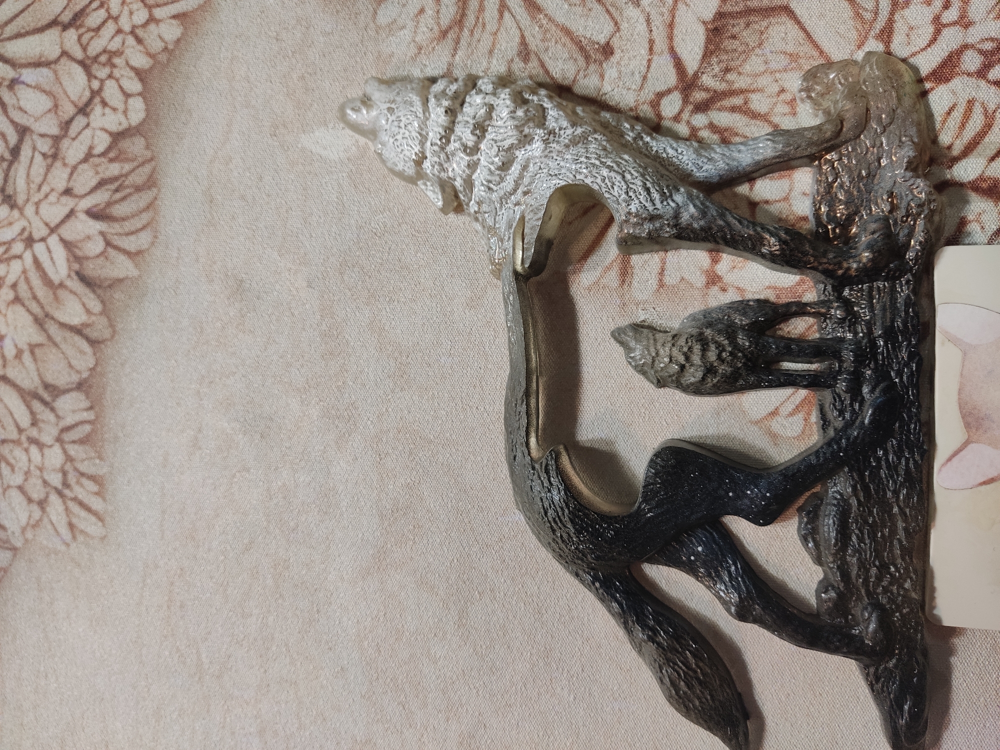 The image shows a close-up of a decorative metal figurine of three wolves on a textured fabric background. The figurine depicts a large wolf on the right, howling with its head tilted back, and a smaller wolf in the center, also howling. A third wolf, larger than the center wolf but smaller than the howling wolf on the right, is positioned on the left, facing left with its head lowered. The wolves are rendered in dark and light metallic colors, with textured detailing to represent fur. The background fabric has a repeating pattern of stylized floral or leaf motifs in shades of brown and beige. The figurine appears to be on a surface that extends slightly out of frame at the bottom, with a sliver of a cream-colored object visible. The scene is static and appears to be an indoor display. The location is not identifiable within the image.